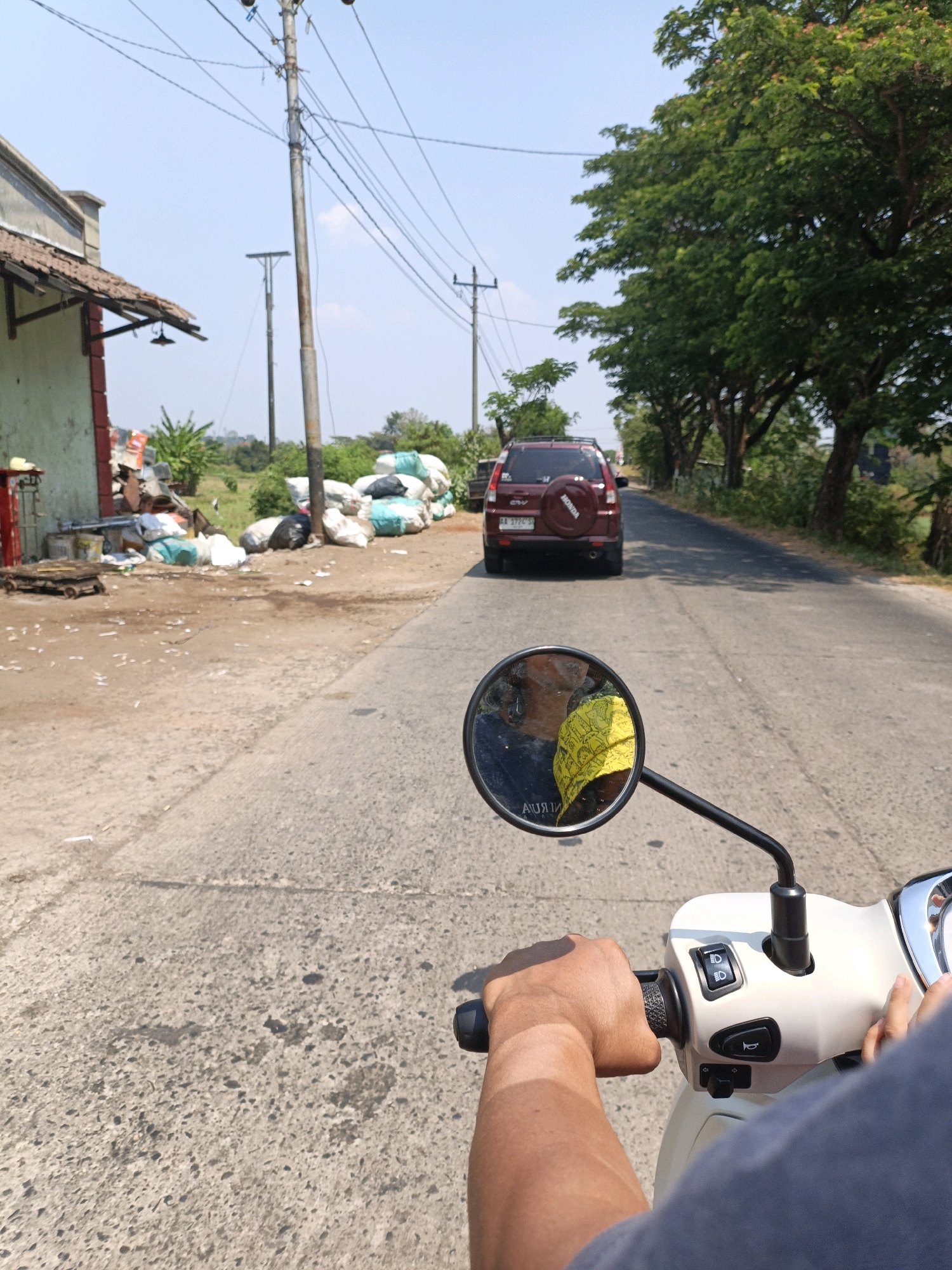 The image is taken from the perspective of a person riding a scooter in Jepara, Indonesia. The scooter's handlebar and a portion of the rider's arm are visible in the foreground. A rear-view mirror on the scooter reflects the rider's face and a yellow cloth. The road ahead is paved and appears to be a rural or semi-urban street, lined with trees on the right side. A red SUV is driving in front of the scooter. On the left side of the road, there is a building with a tiled roof, and a pile of white and green plastic bags, along with other debris. Electrical poles and wires are visible against a clear blue sky. The overall scene depicts a sunny day in a somewhat unkempt area of Jepara, Indonesia.