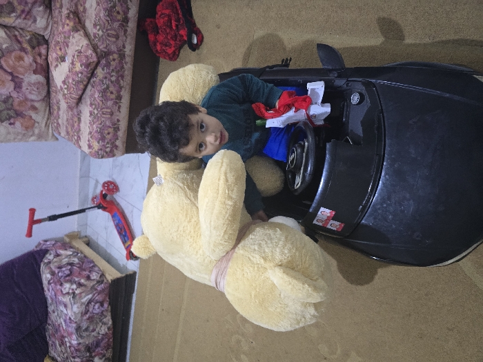 A young child sits in a black toy car, cradled by a large, light yellow teddy bear. The child is wearing a dark green shirt. The car is filled with red and white objects. A red scooter lies on the floor nearby. A floral couch is visible in the background, along with a purple blanket on a different couch. The floor appears to be a light tan color.