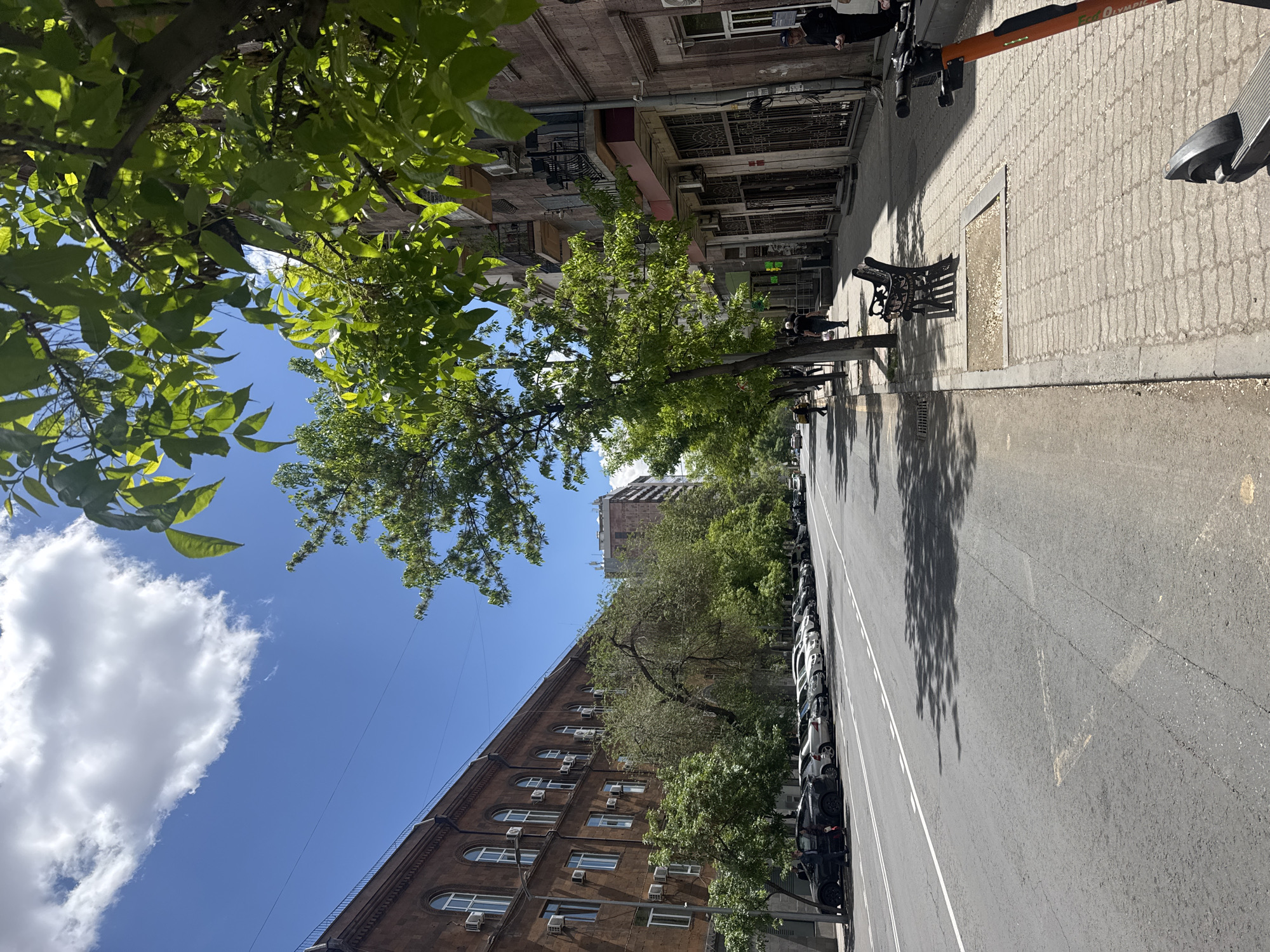 A street scene in Yerevan, Armenia, photographed on a sunny day. The view is along a street with buildings on both sides and a row of trees casting shadows on the road. On the right side of the street is a sidewalk with electric scooters and benches. Cars are parked on the left side of the road, along the building. The sky is blue with some white clouds, visible between the trees. The perspective is slightly tilted.