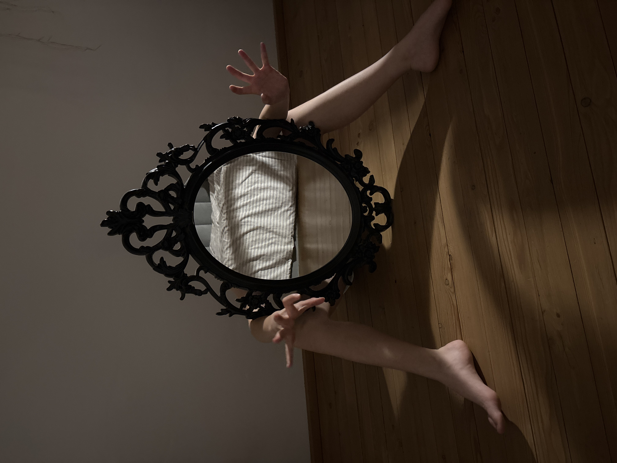 A surreal image shows a person emerging from an ornate, black oval mirror. The mirror is angled diagonally in the frame. The wall behind the mirror is light gray, while the surface behind the person's limbs appears to be a wooden floor. The person's arms and hands extend upward, and legs and feet extend downward from the mirror's frame. The reflection in the mirror is of the person's body and appears to be wearing a white and gray striped shirt. The location, Chaikovsky, Russia, is not visually identifiable in the image.