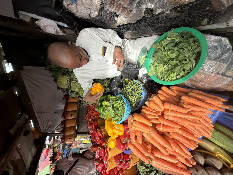 The image depicts an active vegetable market stall in Jakande, Nigeria. In the foreground, a bald man, dressed in a long-sleeved white traditional garment with embroidered details, holds a yellow bell pepper in his left hand and manipulates a black plastic bag with his right, looking downwards at the items. He appears to be a vendor preparing an order.
The stall is abundant with fresh produce. To the man's left and in front of him are large piles of bright orange carrots, bundled with rubber bands, and numerous red and yellow bell peppers. Red bell peppers are also visible in multiple yellow buckets on a shelf behind these. Further back on the shelf are piles of tomatoes. On the counter directly in front of the man are green bell peppers, a blue basket filled with chopped green herbs, and a pile of green beans. To his right, a large green basin holds fresh green leafy vegetables, and a small white bowl contains green peas. Behind these items, stacked cabbages are visible, and to the far right, goods are stored in mesh netting.
In the blurred background to the left, another individual wearing a white t-shirt with a black "ARSENAL" logo is partially visible, facing away from the camera. The market stall has a rudimentary structure with a corrugated roof, wooden supports, and canvas or tarp backdrops. The lighting suggests natural daylight.
