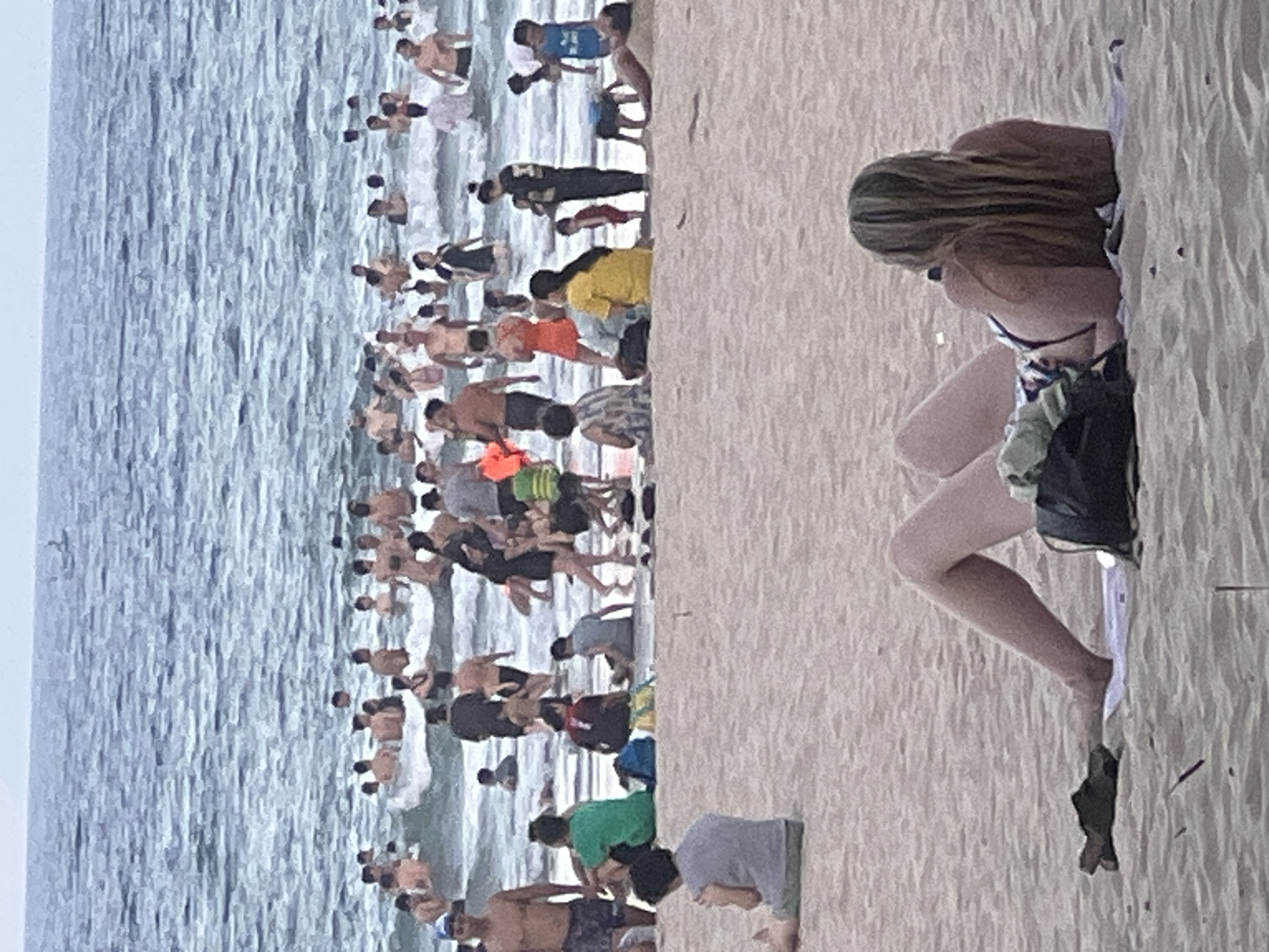 The image depicts a crowded beach scene in Đà Nẵng, Vietnam, with many people enjoying the water and sand. The water appears to be a calm, blue ocean with gentle waves, and the sky is overcast. The sand is light-colored and textured, with footprints and ripples indicating recent activity.
The subjects are predominantly people of various ages, dressed in swimwear and casual beach attire. Many are in the shallow water, some are wading, and others appear to be playing or socializing. On the sand, individuals are seen sitting, lying down, and walking. A woman in a bikini is prominently featured in the foreground, lying on a towel on the sand.
The activity is clearly recreational, with people enjoying a day at the beach. The atmosphere seems relaxed and casual, typical of a summer day at the seaside. The time of day is difficult to ascertain definitively due to the overcast sky, but the lighting suggests it could be mid-morning or afternoon.
Notable details include the density of the crowd, indicating a popular beach destination. The presence of beach towels and bags suggests people are settling in for an extended stay. The overall scene is one of community enjoyment and leisure. There is no visible text in the image.