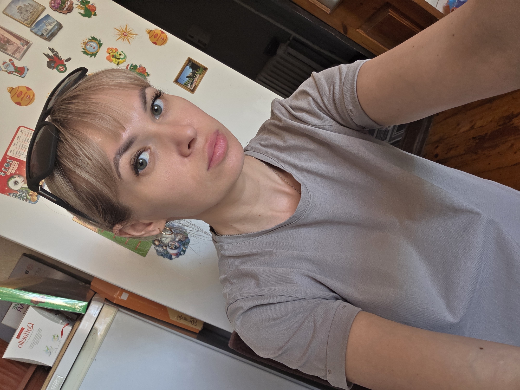 A selfie of a blonde woman with a slightly quizzical expression. She has sunglasses perched on her head. Behind her appears to be a refrigerator covered in magnets, and a wooden cabinet. Based on some of the magnets, she may be in Chaikovsky, Russia. She is wearing a gray t-shirt.