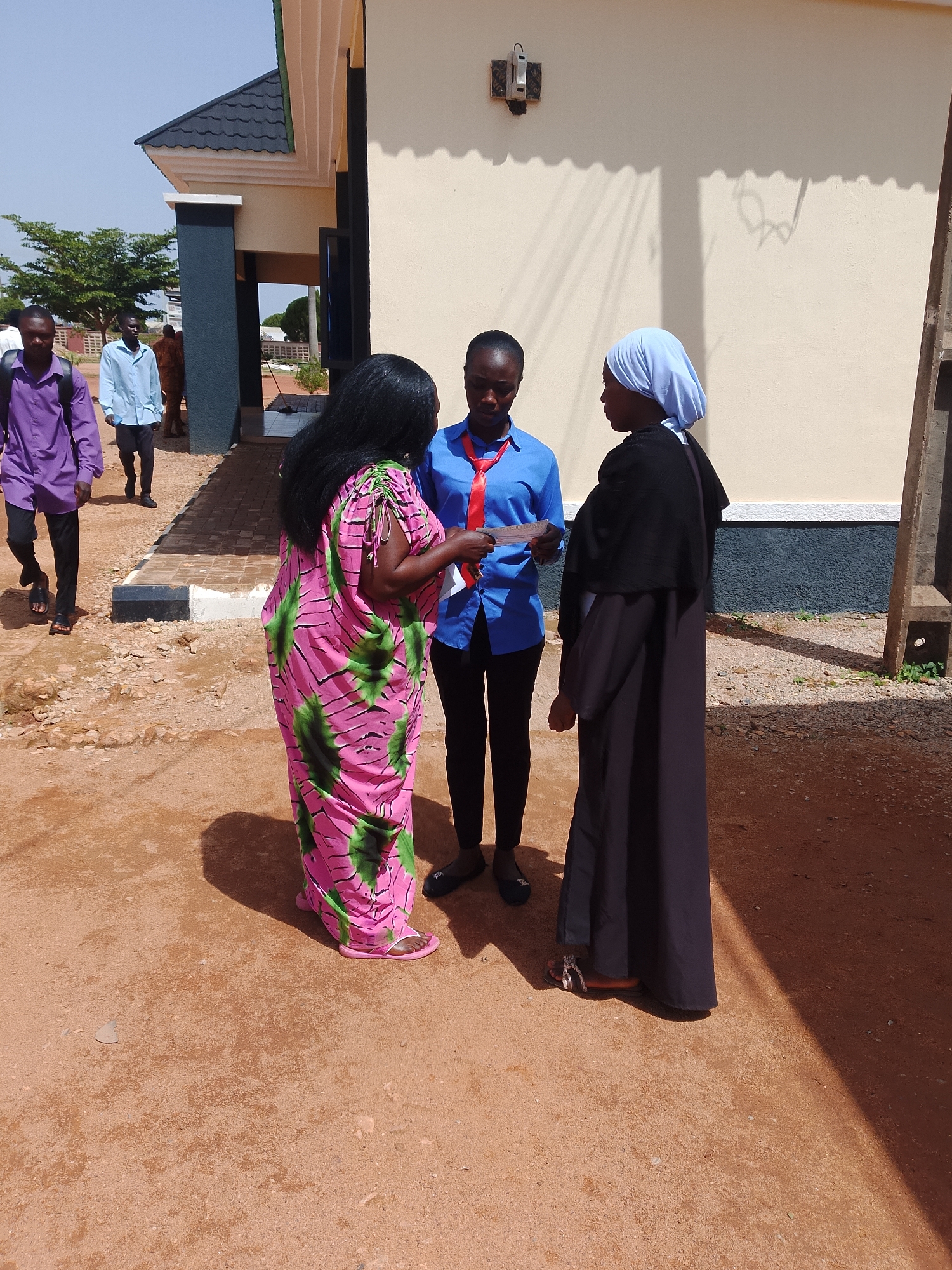 In Jos, Nigeria, three people are engaged in a discussion outdoors in front of a building. A woman wearing a vibrant pink and green patterned dress stands on the left, facing two younger individuals. To her right, a young woman in a blue uniform with a red tie looks down, holding a piece of paper. Beside her, a woman in a black abaya and a light blue headscarf also holds a document and appears to be listening. The ground is covered in reddish-brown dirt, with some scattered pebbles. In the background, part of a light-colored building with dark trim is visible, as well as trees and a clear sky. Several other individuals are seen walking in the background. The scene suggests a casual outdoor meeting or interaction.
