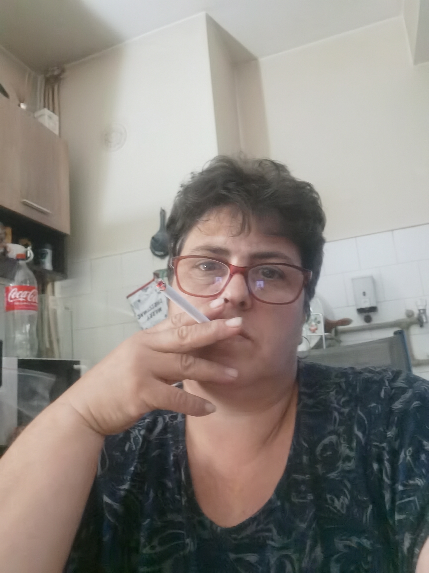 An indoor, medium-shot, selfie-style photograph shows a person with short, dark, curly hair and red-framed glasses. The person is holding a lit cigarette in their right hand, near their mouth, with smoke obscuring part of their face. They are wearing a dark blue t-shirt with a light grey, swirling pattern. In the background, a white tiled wall is visible, with some kitchen cabinets to the left. A bottle of Coca-Cola is on a shelf in the cabinets. The environment appears to be a kitchen or similar domestic setting. The location is not explicitly identifiable as Lomets, Bulgaria, based solely on the visual information present.