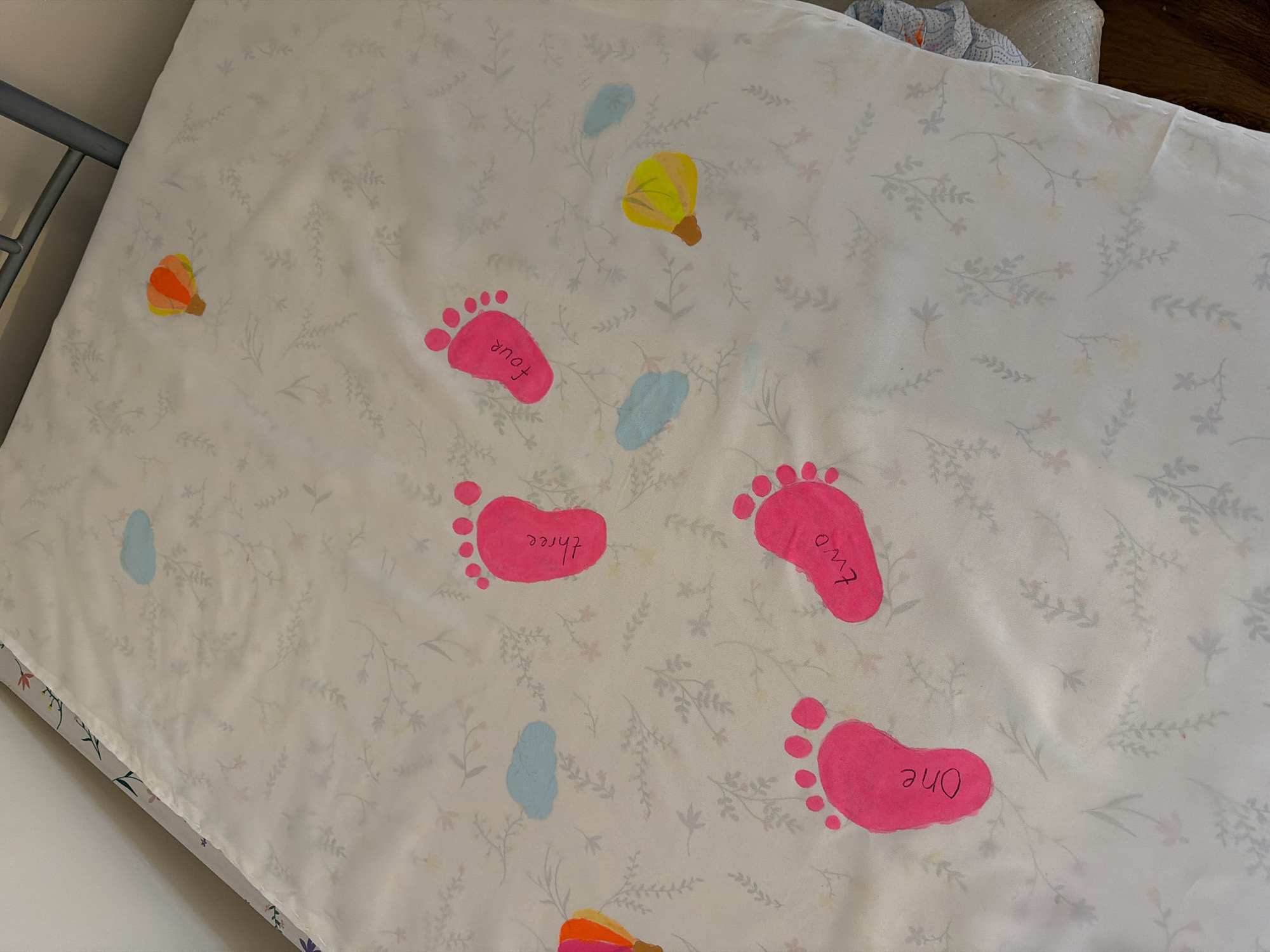 A white mattress pad with a floral design rests on a mattress and frame. Scattered across the pad are cartoonish depictions of bare footprints, colored hot pink and each labeled with a number in handwritten text: "one", "two", "three", and "four." Also on the pad are small, stylized drawings of hot air balloons, colored yellow/brown and orange/brown, along with three small blue shapes that could represent raindrops or blobs. The scene's location is unspecified and unidentifiable in Bearpark, United Kingdom.