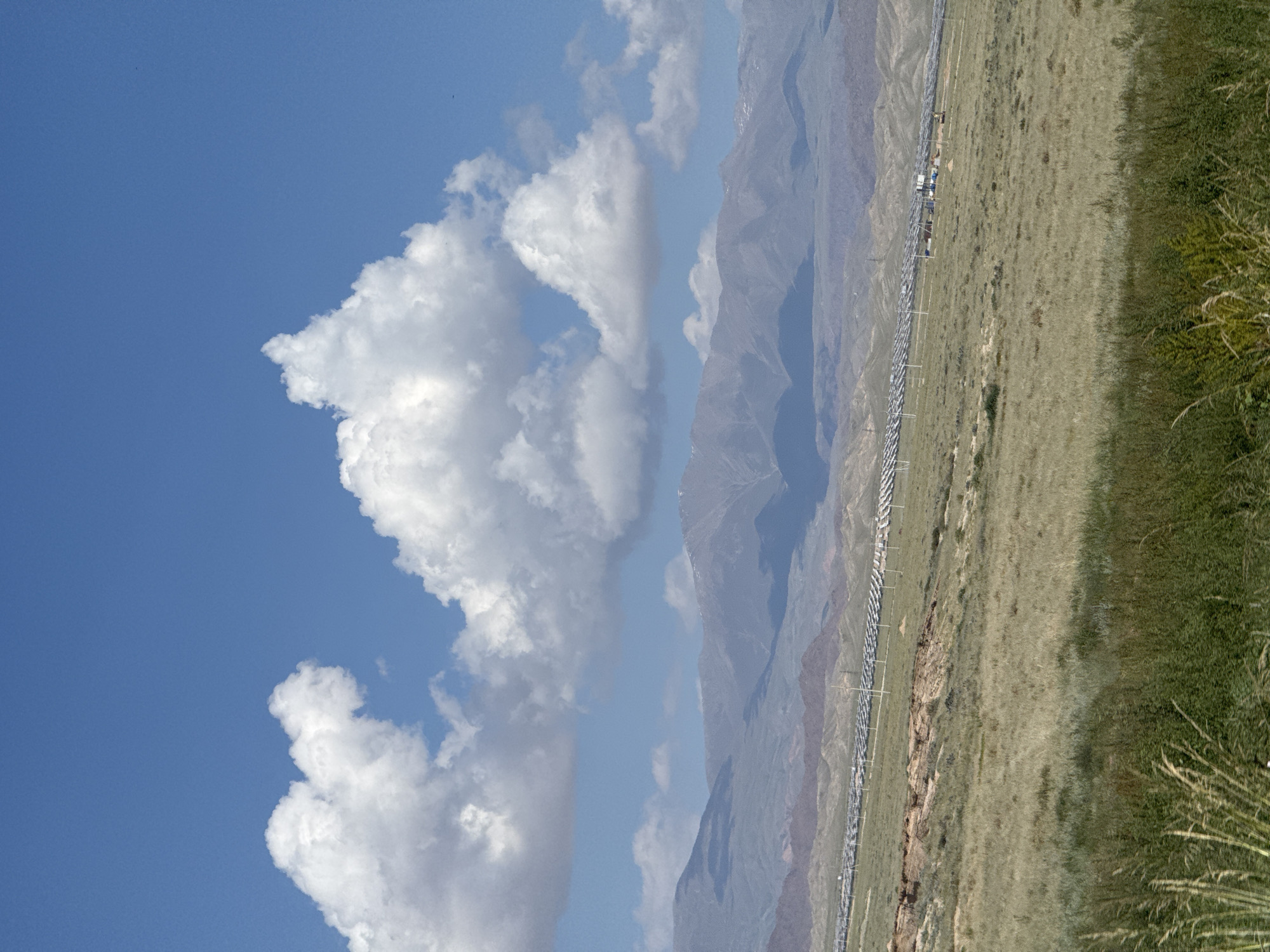 The image captures a wide-angle landscape view under a bright blue sky with fluffy white clouds. In the foreground, a grassy hillside slopes gently downwards. In the middle ground, a large array of solar panels is visible, stretching across the landscape. Beyond the solar panels, a series of rolling hills and mountains form the background, with some appearing to have snow-capped peaks. The scene suggests an open, semi-arid environment, characteristic of the Toru-Aigyr region in Kyrgyzstan, known for its vast open spaces and increasing adoption of solar energy. There are no people or specific actions depicted, just the static environment.