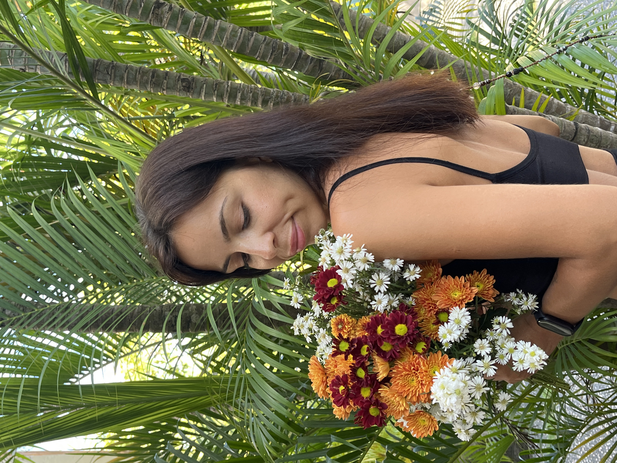 The image features a young woman positioned among lush green palm trees, consistent with a tropical setting like Kuta Selatan, Indonesia. The woman, with long, dark brown hair and light-brown skin, is seen from a side-back angle, with her head turned to her left. She is wearing a black spaghetti strap top and has a dark-colored smartwatch on her left wrist.
She is holding a vibrant bouquet of flowers, primarily composed of white, orange, and deep red daisy-like blooms, likely chrysanthemums. Her head is tilted towards the flowers, and she has a gentle, serene expression with a soft smile, eyes closed or looking downwards, suggesting a moment of quiet contentment or appreciation for the flowers.
The background is dominated by the intricate patterns of green palm fronds and the textured trunks of palm trees, indicating an outdoor, daytime scene with ample natural light. There are no visible texts or specific weather conditions apparent beyond clear, bright sunlight.