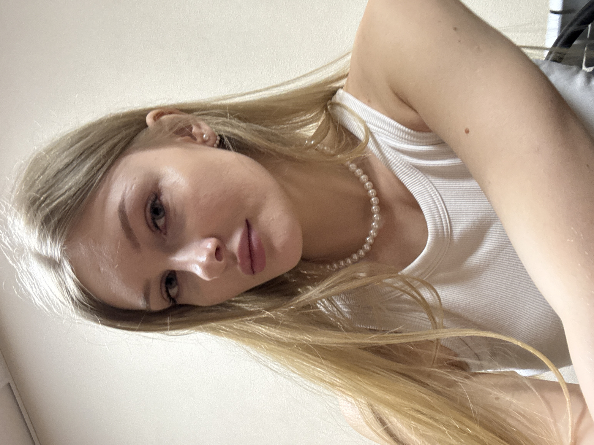 A fair-skinned young woman with long blonde hair is positioned in the center of the frame. She is wearing a white tank top and a pearl necklace. Her head is tilted to the left, with her face angled towards the camera. Her expression is neutral, with light makeup on her lips. The background is a plain off-white wall. No location information about Perm, Russia is present.