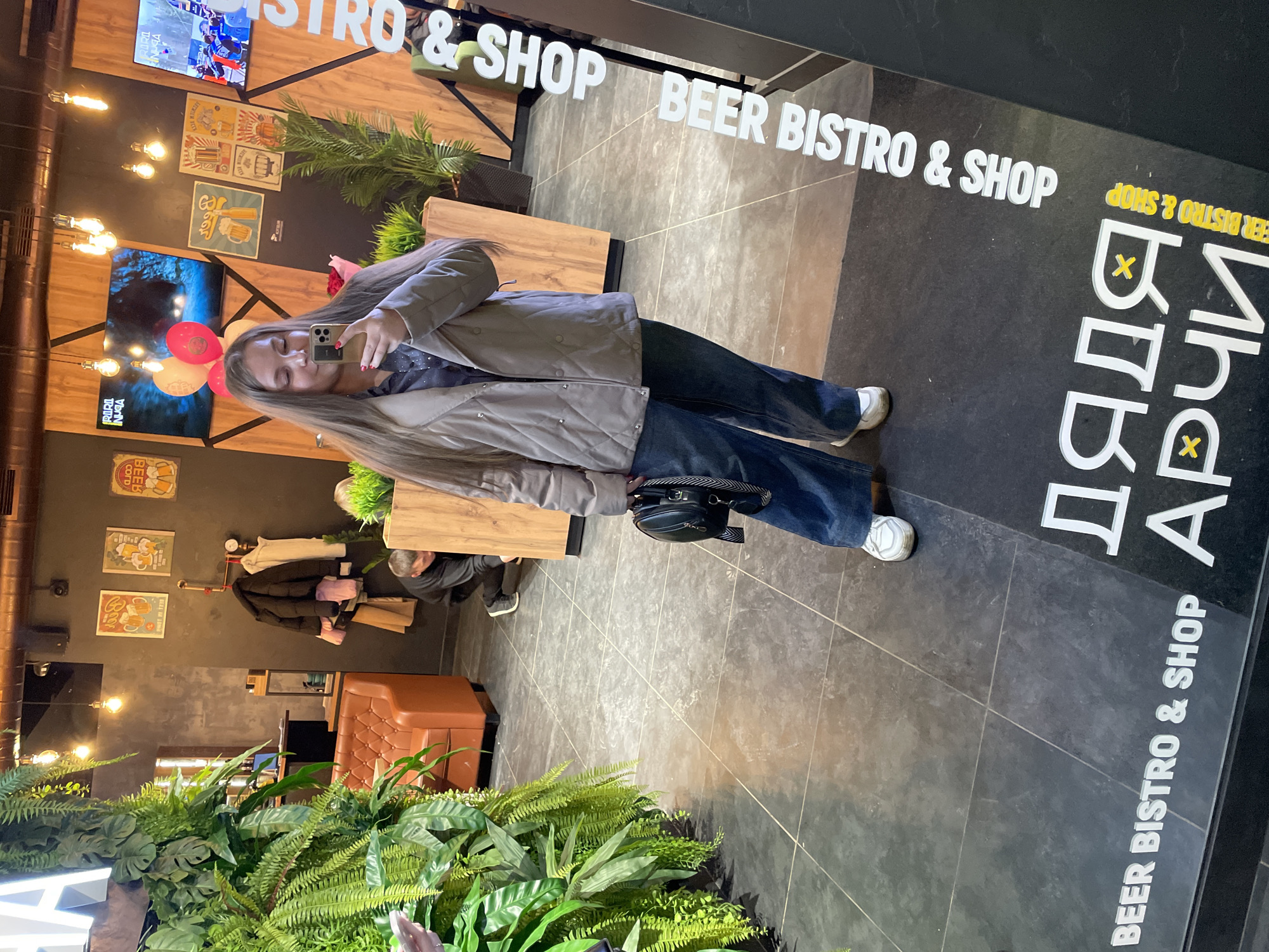 A young woman with long blonde hair takes a selfie in a commercial establishment identified as "Beer Bistro & Shop" in Izhevsk, Russia. She is wearing a quilted jacket and jeans, holding her phone with a brown case. The establishment is decorated with plants, and displays posters and TVs on the wall. The name "ДЯДЯ АРЧИ" (Uncle Archie) is printed on the floor. A person is kneeling down in the background, possibly cleaning or stocking the store.