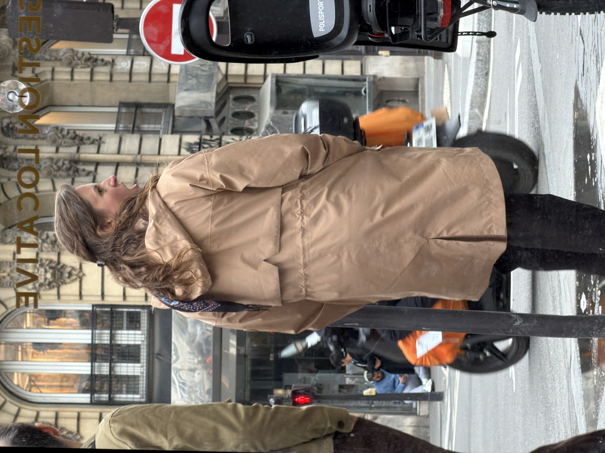The image shows a woman with long brown hair wearing a tan coat and black pants. She is walking on a street in Paris, France. In the background, there is a building with large windows and decorative architecture. The words "DESTINATION LOCATION" are vertically written along the side of the building. A moped is parked nearby, with its front wheel visible on the street. Other people are visible in the distance. The scene seems to be a snapshot of everyday urban life in Paris.
