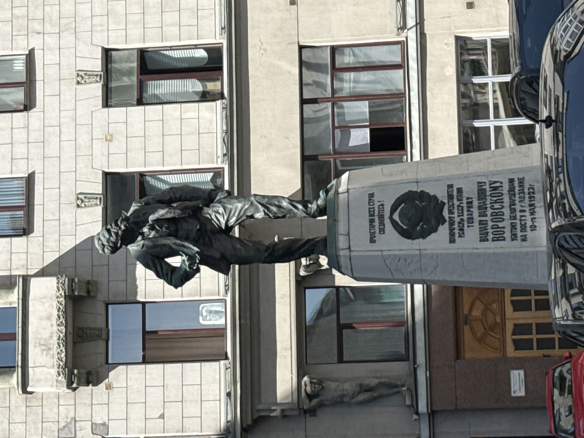 The image shows a monument in Moscow, Russia, dedicated to Václav Vorovský. A bronze statue of a man in mid-stride is situated next to a stone plinth. A portrait of Václav Vorovský is inscribed on the plinth, as is text in Russian. The building in the background has a facade that is a grid of window bays and stone blocks. The lower ground floor of the building is marked by a sculpture of a reclining figure. A black vehicle is visible to the right of the monument, and a red vehicle is visible towards the bottom right corner.