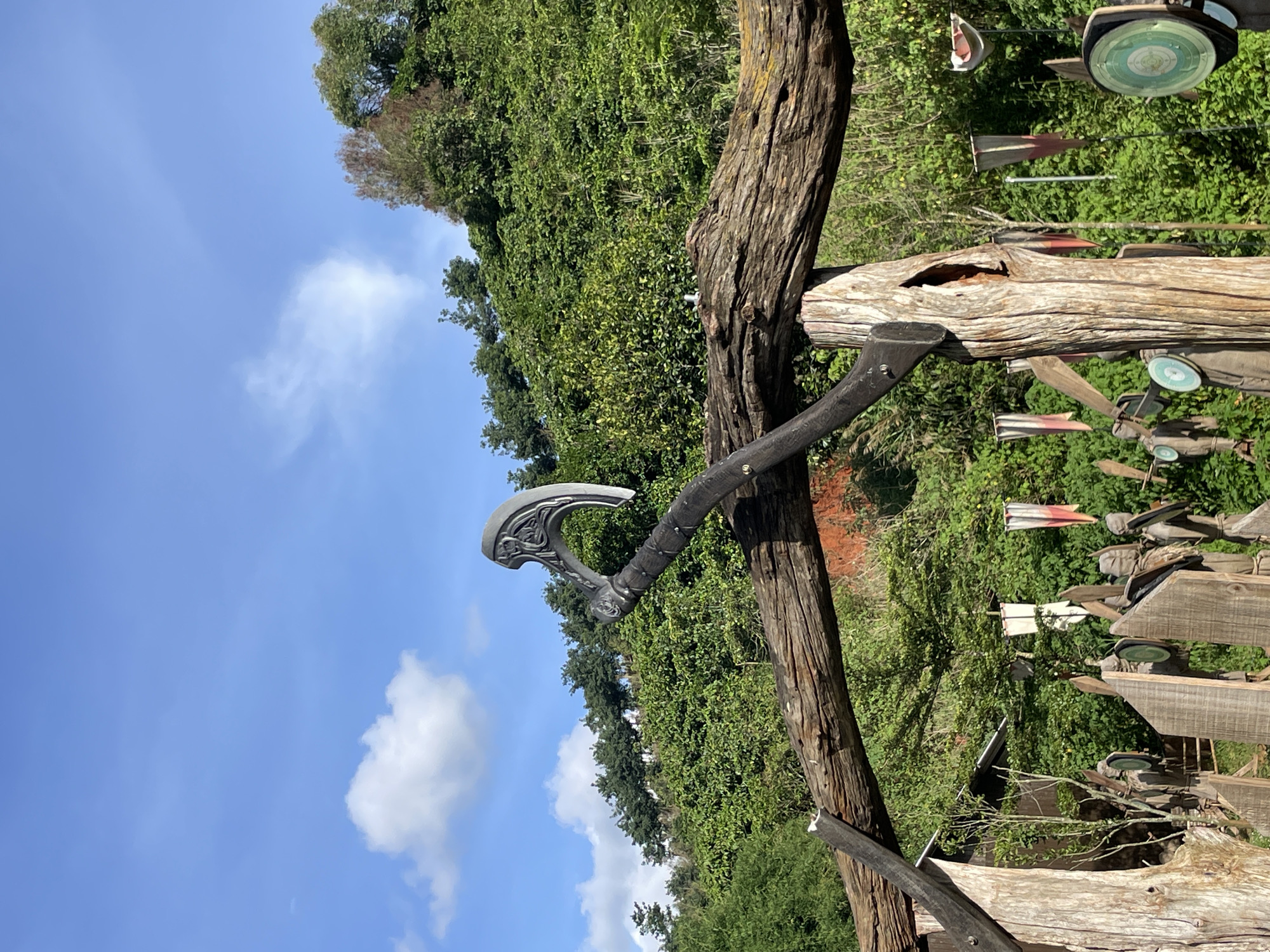 An elaborate, prop axe rests on a weathered wooden archway in Thôn 1, Vietnam. The axe, with a decorative, crescent-shaped blade, is propped on a thick, gnarled log that forms the horizontal beam of the arch. The arch itself is constructed from rough, unfinished logs. Behind the arch, a lush, green hillside is densely covered in vegetation, with a clear blue sky overhead dotted with a few white clouds. Below the arch, wooden figures that resemble warriors, some adorned with flags and circular shields, stand in a line. The scene appears to be an outdoor display or attraction.