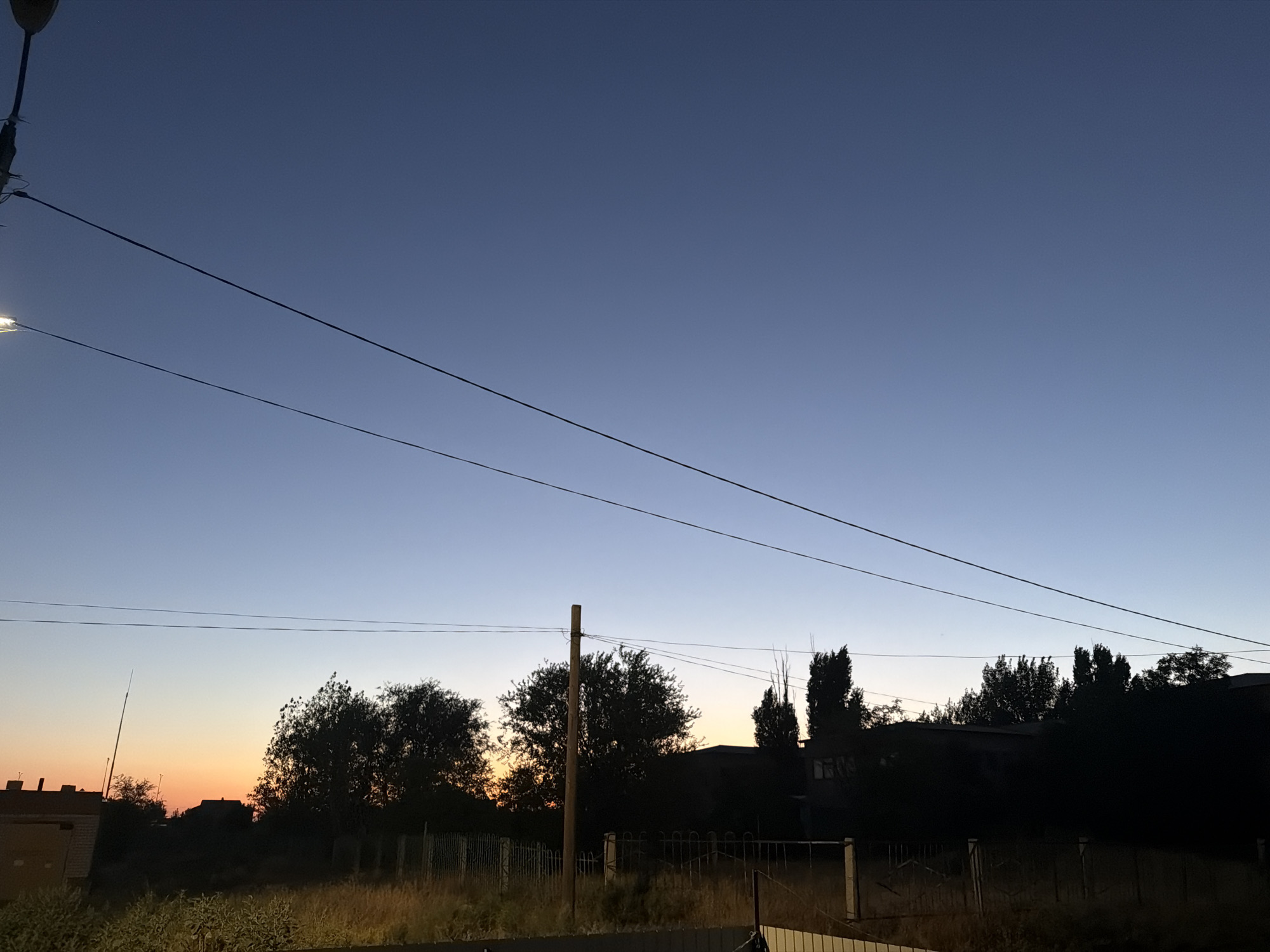 The image depicts a twilight scene in the village of Malyye Derbety, Russia. The sky transitions from a deep blue at the top to a pale orange near the horizon, indicating either sunrise or sunset. Silhouetted against the sky are trees, fences, and the rooftops of several buildings, suggesting a residential or semi-rural area. Power lines are prominently stretched across the frame, dividing the sky. In the foreground, dry grass and vegetation are visible. The overall impression is one of a quiet, serene evening.