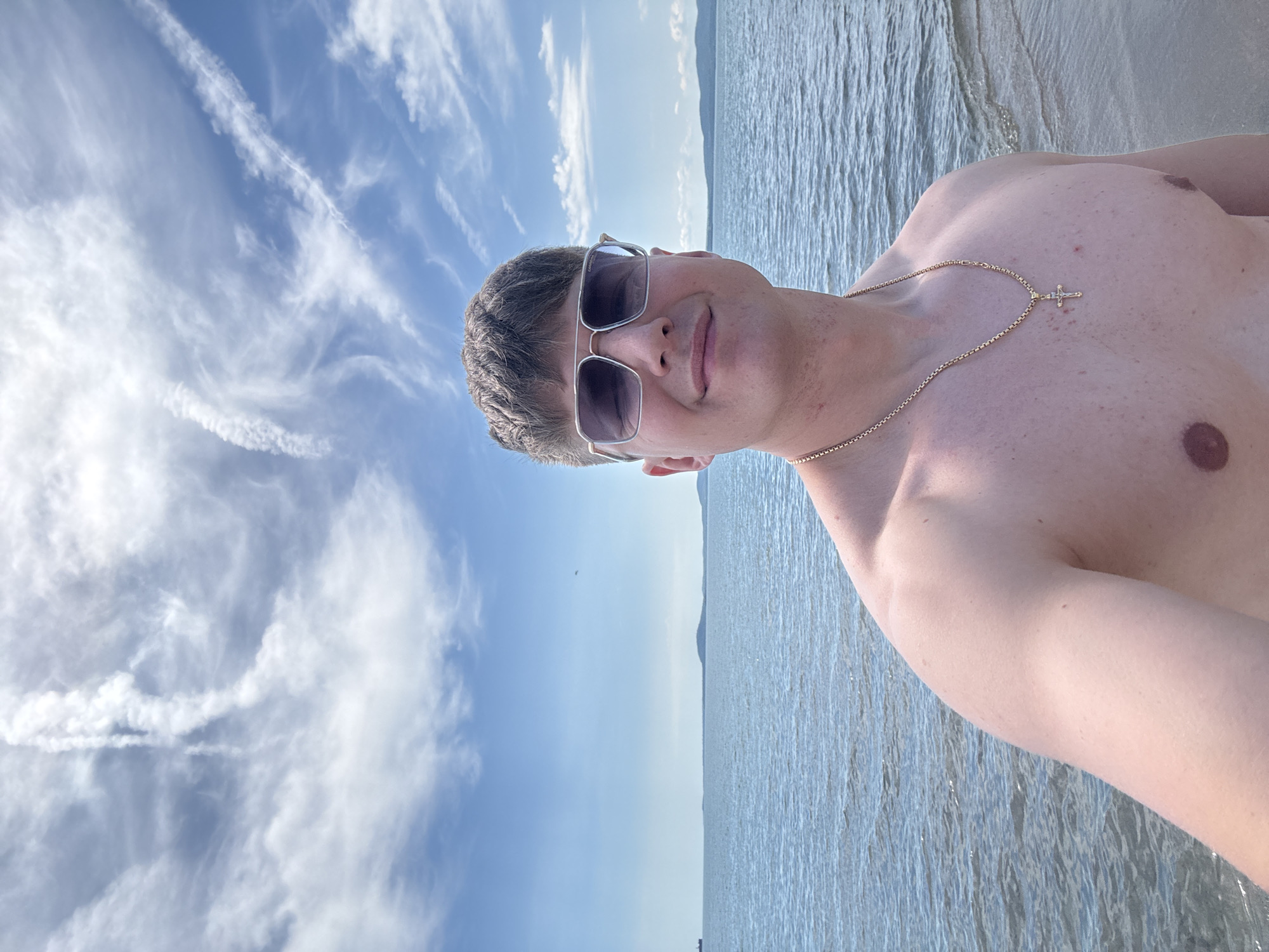 A slightly-rotated selfie shows a light-skinned person wearing sunglasses and a gold necklace with a cross pendant. The person is shirtless and positioned in front of water and sky. The sky is light blue with scattered, streaky clouds. A faint line of land is visible in the distance where the sky meets the water. The photo was likely taken in Burgas, Bulgaria, given the location information.