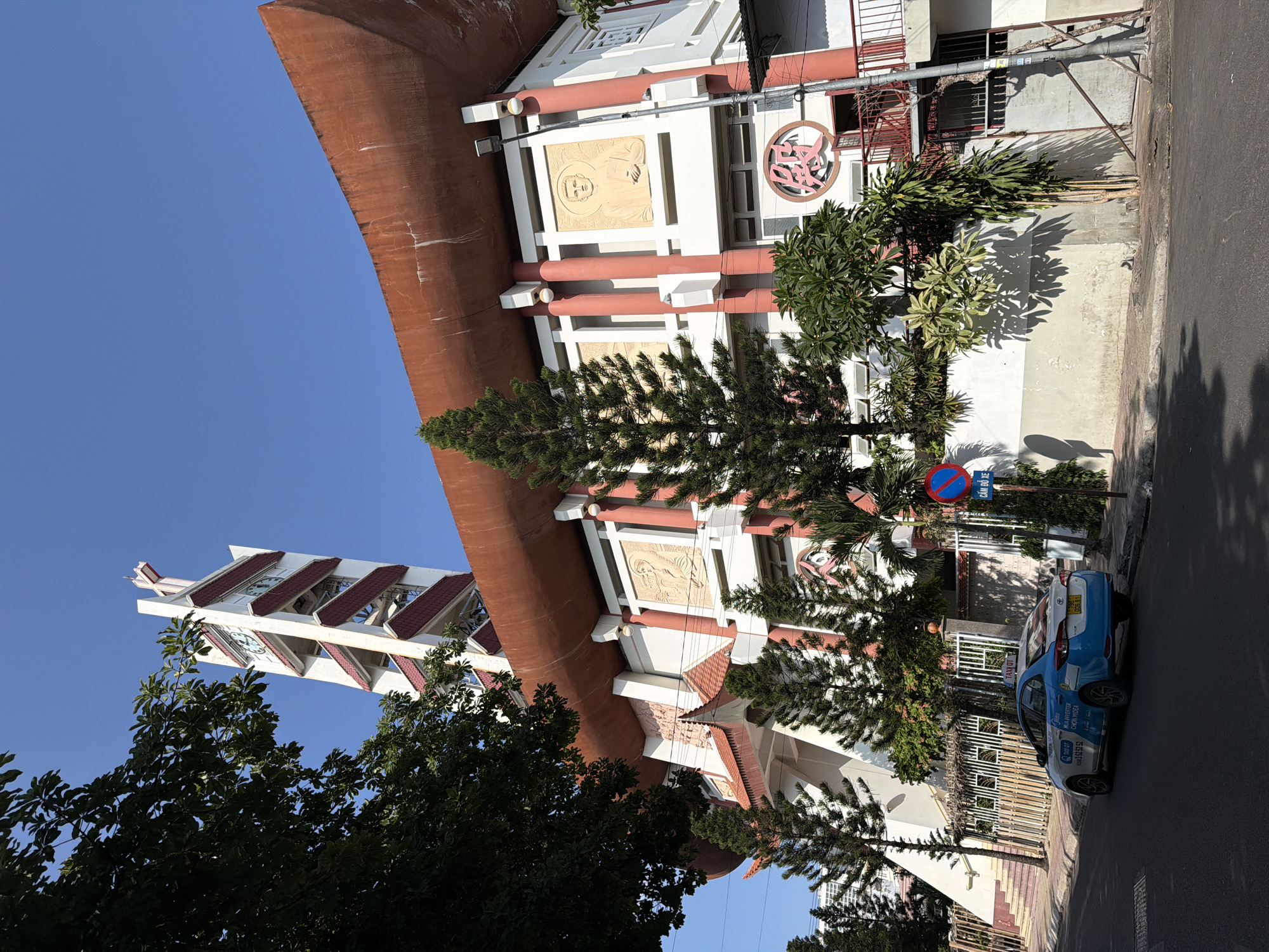 The image shows a wide-angle, low-angle view of the Tan Dinh Church in Nha Trang, Vietnam. The church, a prominent landmark, features a distinctive bell tower with a white facade and red accents, topped with a cross. The main church building has a large, curved, reddish-orange roof and white walls with red pillars. Relief carvings of figures are visible on the facade, and a large circular emblem with Vietnamese characters is also present. In the foreground, a blue and white taxi with "TAXOT" branding is parked on the street. A street sign with red and blue markings and Vietnamese text is posted on the sidewalk. Lush green trees and foliage surround the church. The sky is a clear, bright blue. The scene is captured on a sunny day, with shadows cast on the ground and the building.