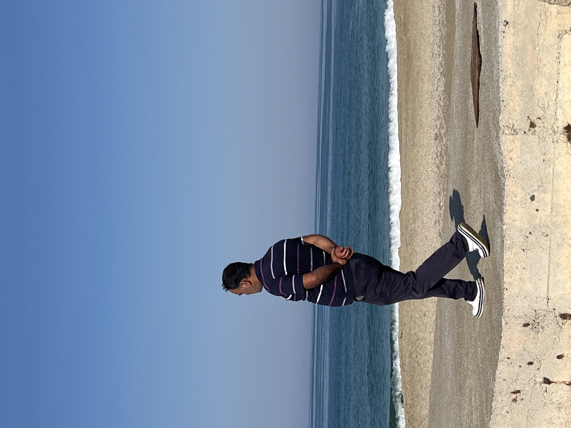 A medium shot, taken at an oblique angle, depicts a man standing on a concrete structure adjacent to a beach, potentially in Alanya, Turkey. The man, facing left, has his hands clasped behind his back. He is wearing a horizontally striped, collared shirt and dark-colored pants, along with white-and-black shoes. The ocean is visible in the background, with white-capped waves meeting the sandy beach. The sky is clear and blue. The concrete structure appears aged with some discoloration. A partial shadow cast by the man is visible on the concrete.