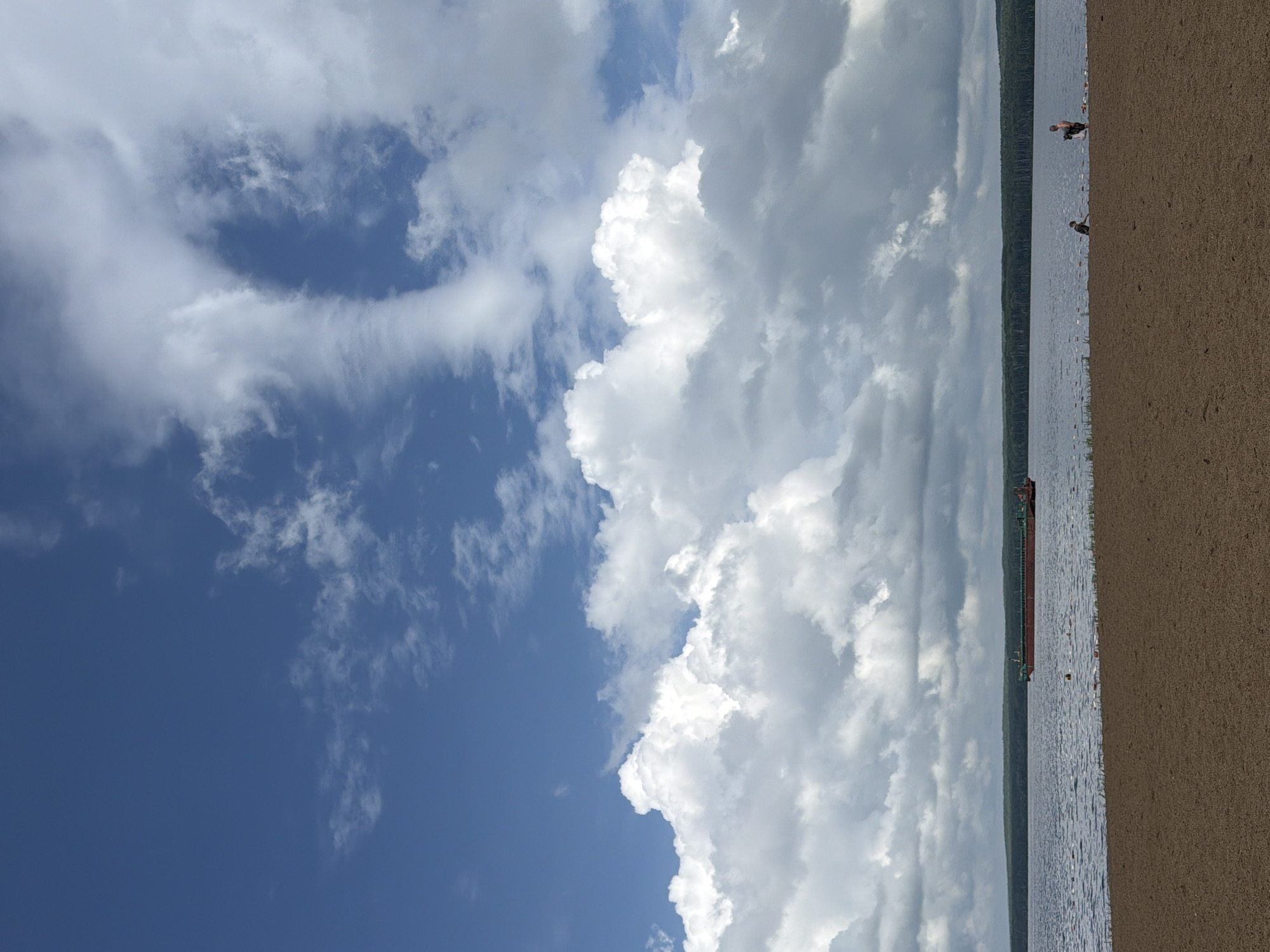 This media file depicts a beach scene on a body of water. The image is oriented vertically. On the left side, a clear blue sky transitions into a vast expanse of white, puffy clouds that dominate the upper portion of the frame. On the right side, a sandy beach occupies the foreground, leading to a calm body of water. The water stretches horizontally across the image, with a distant tree line forming a dark green border in the background.
Several small figures are visible near the water's edge. One figure, a person, stands in the shallow water, while another, possibly a child, is crouched on the sand near the water. Further out on the water, a long, dark barge is moving parallel to the shore. Buoys are spaced out in the water, possibly marking a swimming area.
The lighting suggests it is daytime, with sunlight filtering through the clouds, creating a somewhat diffused illumination. The weather appears partly cloudy, with dramatic cloud formations. The presence of a beach and the barge suggest a recreational or industrial waterfront setting, potentially a lake or a wide river. The scene evokes a sense of leisure and activity on a summer day. No visible text is present in the image.
