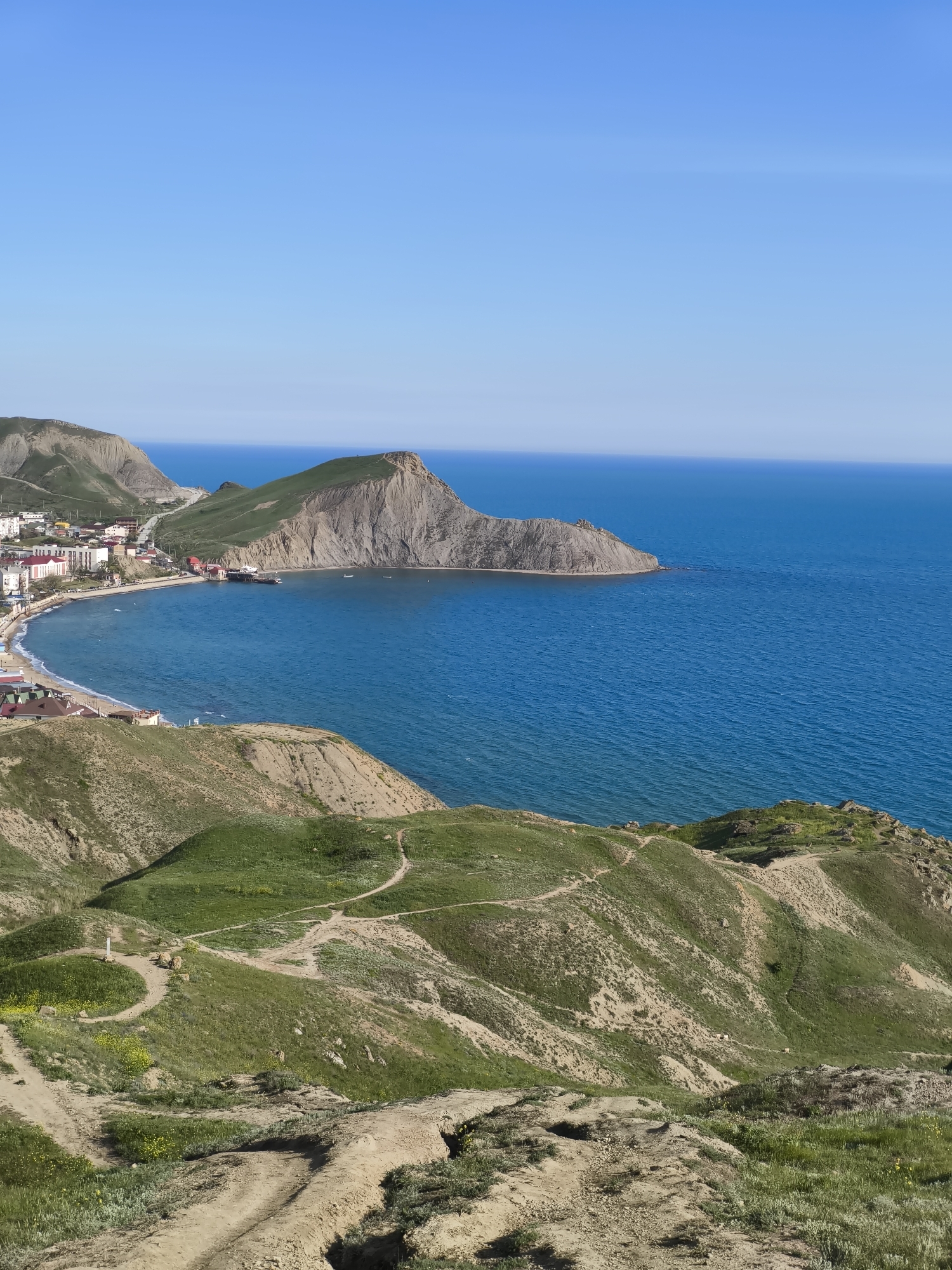 The image shows a coastal landscape under a clear blue sky. The foreground features a series of hills covered with patches of green vegetation and dirt paths. In the middle ground, a town sits along the coast of a bay, with buildings clustered near the beach. A rocky headland extends into the blue sea, forming a prominent point. The water is relatively calm, and the sky is clear. The location appears to be in or near the city of Unknown, Unknown.