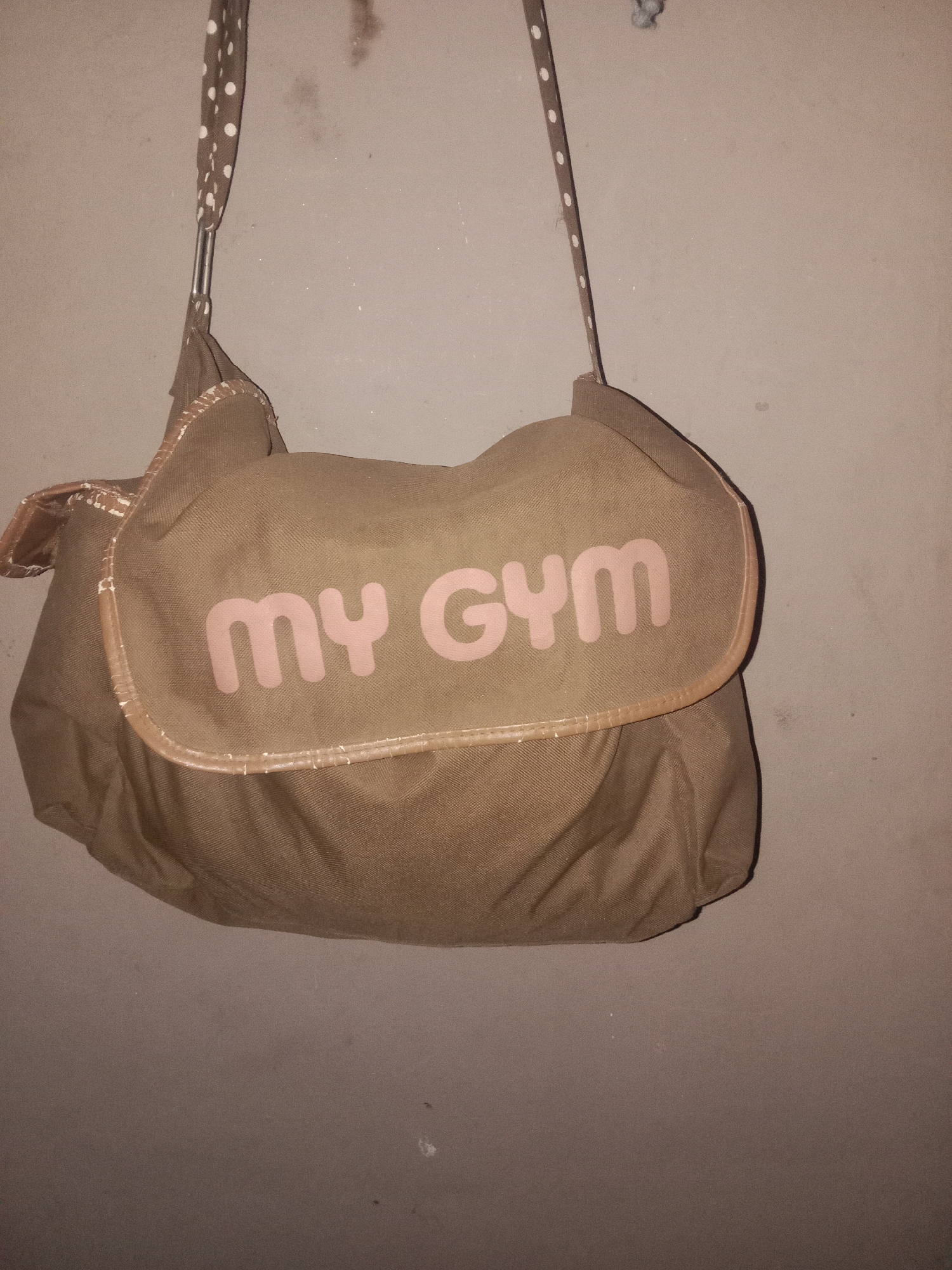 A brown messenger bag with pink lettering that reads "MY GYM" is hanging on a light brown wall. The bag has a flap closure with a lighter brown trim and a polka dot strap. The wall appears to be made of concrete or stucco and has some smudges and dust on it. The lighting is dim, casting shadows on the wall and bag. The bag is the primary subject of the image.