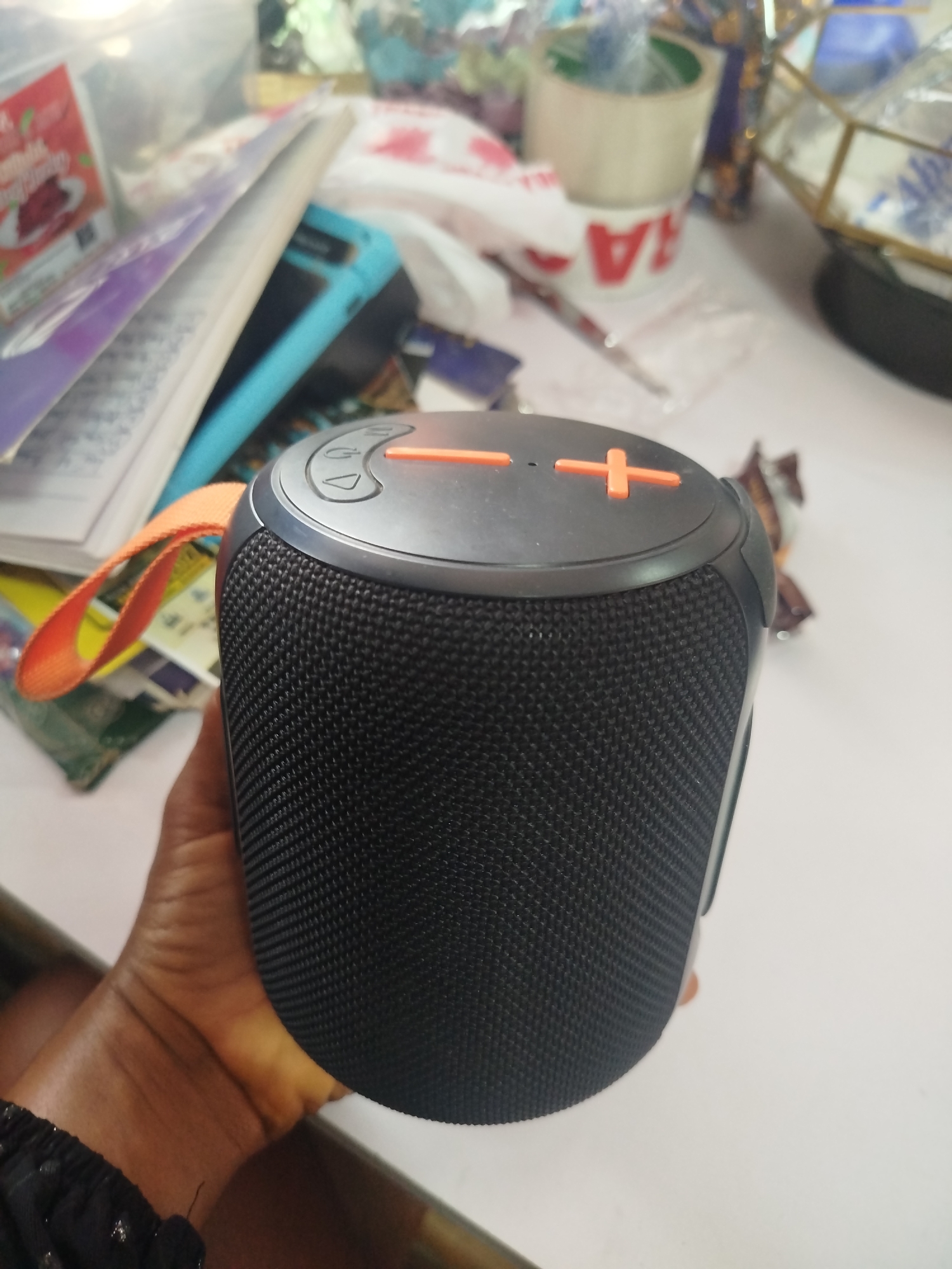 In Ugbowo, Nigeria, on a white surface, a dark grey, cylindrical speaker is held by a person's hand. The speaker has a textured black fabric grill and a silver top with orange plus and minus buttons. An orange fabric loop is attached to the side of the speaker. In the background, various items are scattered, including books, a blue phone case, rolls of tape, and a clear glass geometric object. The scene appears to be indoors, on a table.