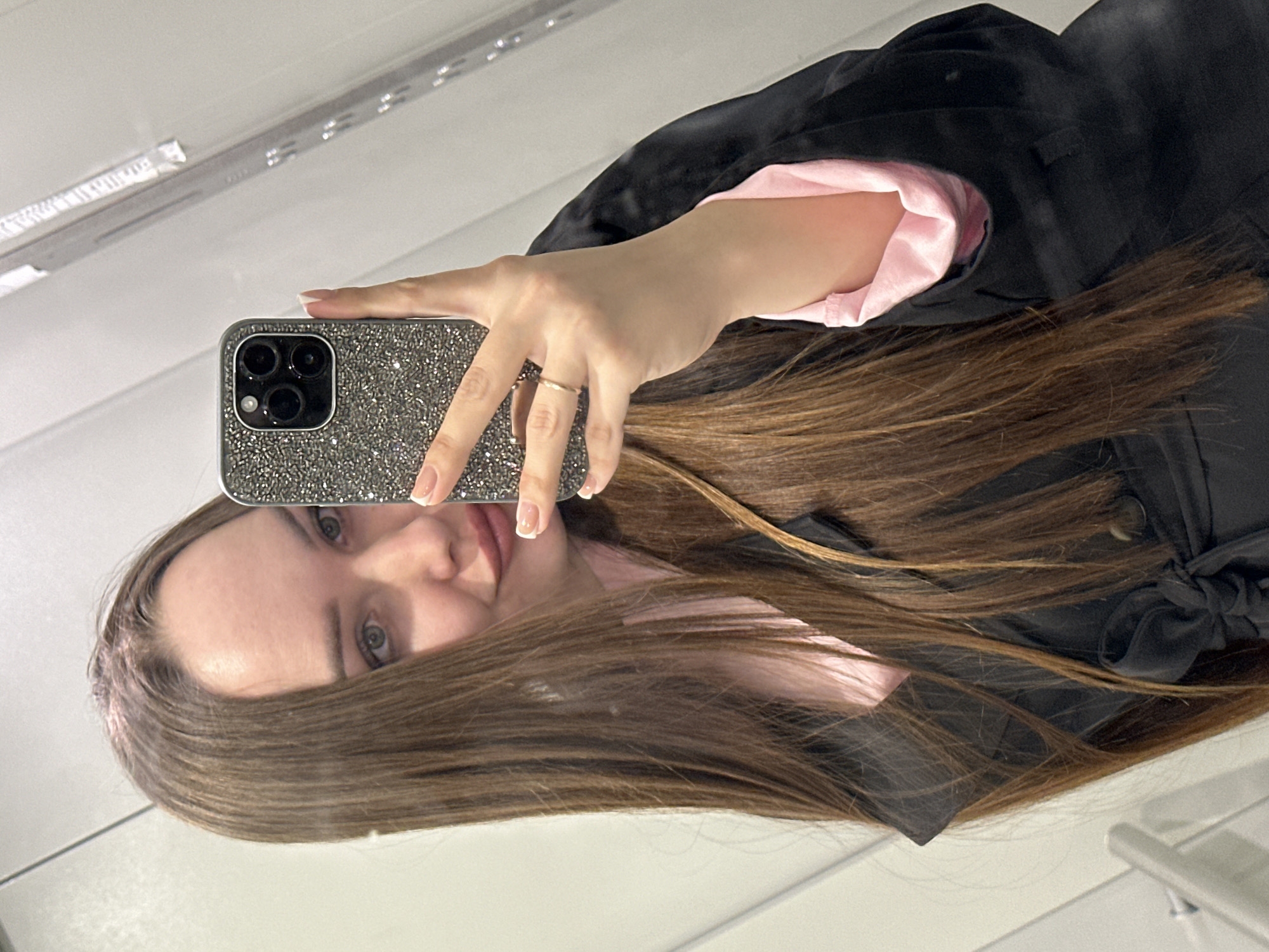 The image displays a female individual taking a mirror selfie in an indoor setting within the city of Chaikovsky, Russia. The subject, visible from the chest upwards, has long, straight, brown hair and light-colored eyes, with a slight smile evident. She is wearing a black coat, possibly a trench coat, tied at the waist, over a light pink garment. Her right hand, featuring French-manicured nails and a thin gold-colored ring on the ring finger, holds a smartphone with a dark, glitter-embellished case and a triple-lens camera array. The reflective surface serving as the background exhibits vertical panel lines, with a metallic handle visible in the lower left reflection and a light fixture or control panel element reflected in the upper right.