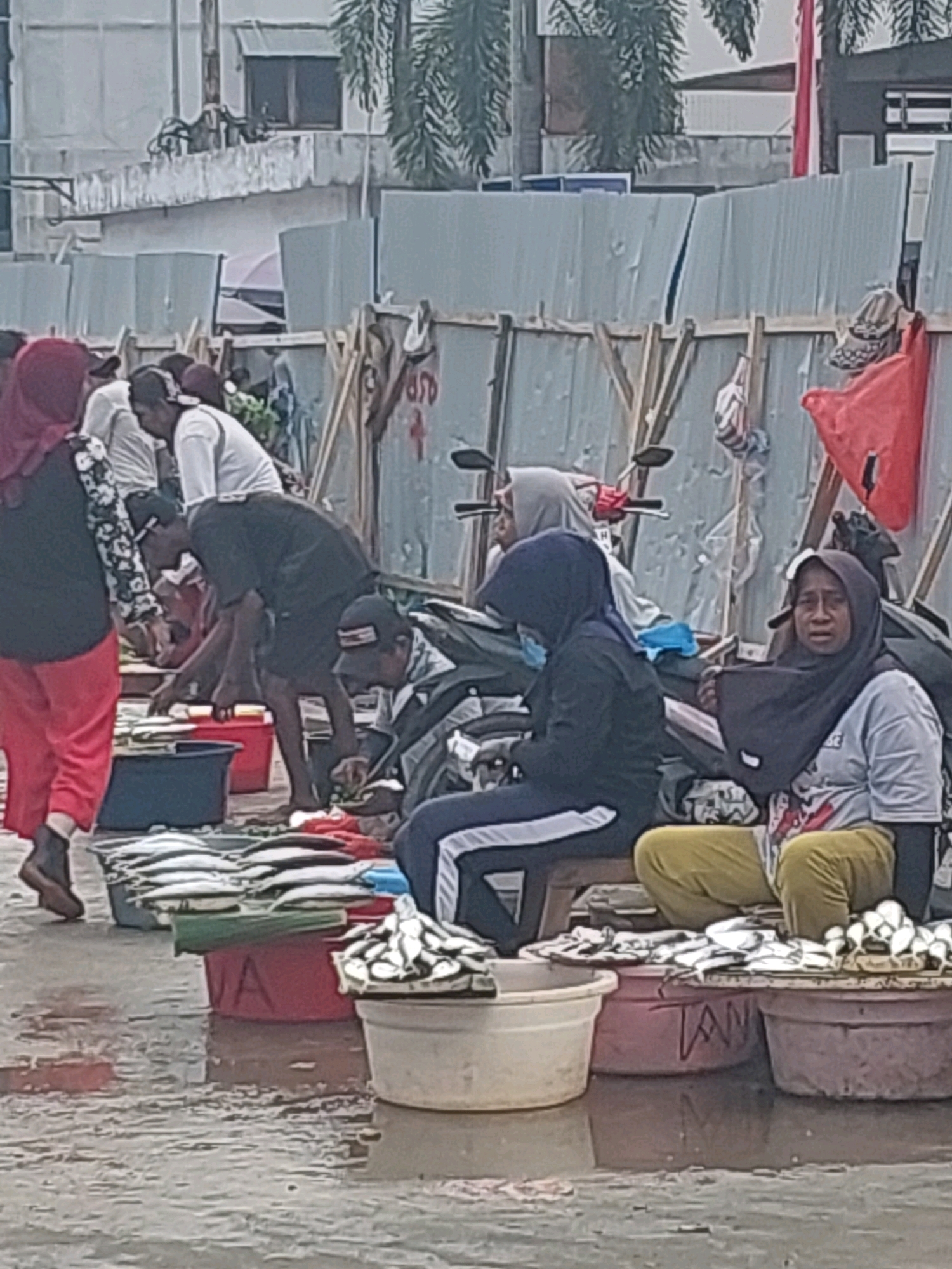 The image depicts an outdoor market scene in Ambon, Indonesia. Several individuals are present, some of whom are selling or arranging fish displayed in various containers. In the foreground, large buckets and tubs hold piles of fresh fish. The ground is wet, suggesting recent rain or the market's proximity to water. In the background, makeshift stalls constructed from wood and corrugated metal panels line the market area. The overall environment appears to be a bustling, somewhat rustic, and functional marketplace.