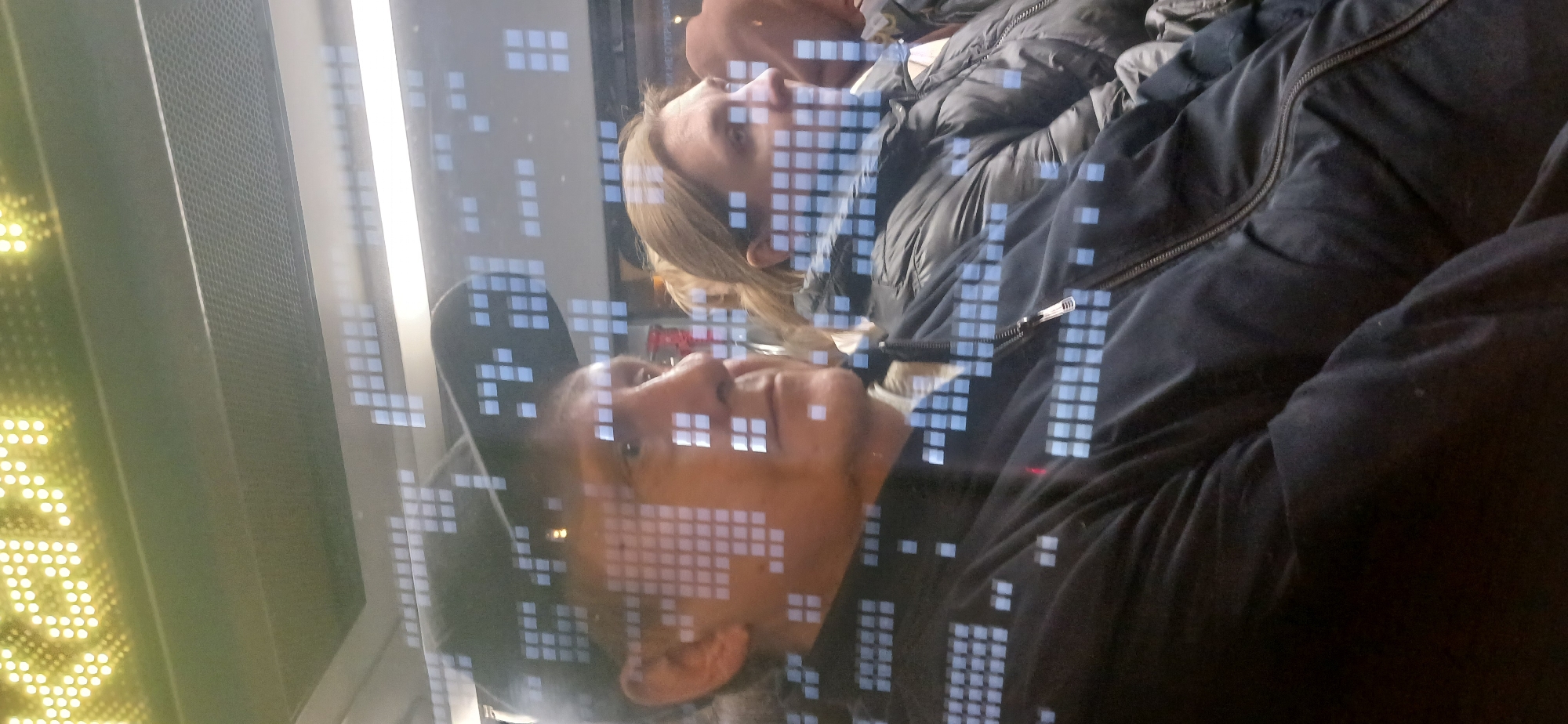 The image shows a reflection of two people, a man and a woman, in a window. The man is wearing a black jacket and a black baseball cap. The woman has blonde hair and is wearing a light-colored coat or jacket. The window also reflects a digital display with a pattern of illuminated squares, possibly a train schedule or advertising, which suggests the scene is likely set on a public transit vehicle like a train or bus. Given the context of Moscow, Russia, it could be a Moscow Metro train car or a similar transit system displaying information.