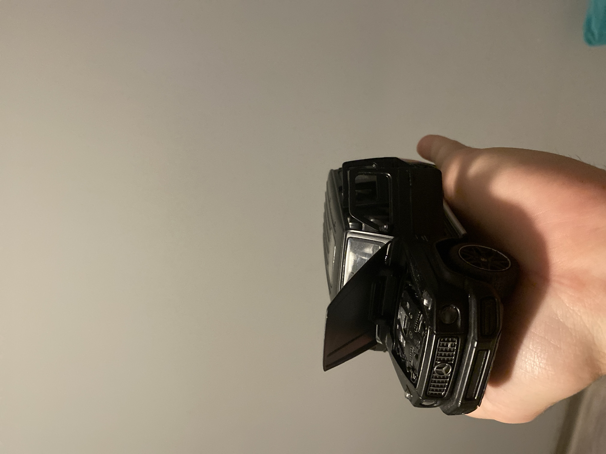 A black toy Mercedes-Benz G-Class is held in a person's left hand. The toy car's hood and driver's side door are open. The background is a plain, light-colored wall. The image does not contain any identifiable landmarks or clues to determine its location in Łódź, Poland.