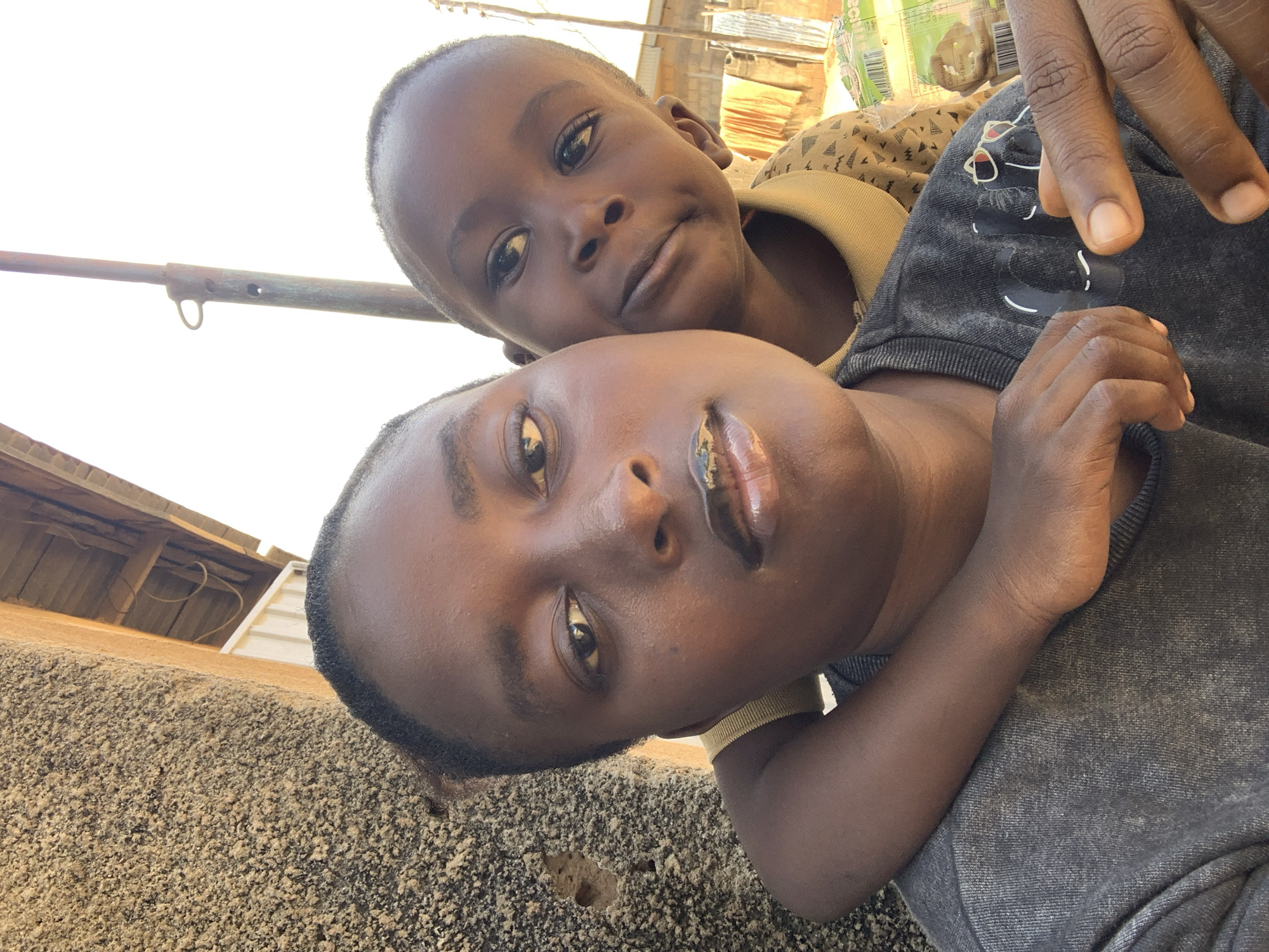 A close-up selfie shows two individuals, a woman and a young child, in an outdoor urban setting. The woman is in the foreground, with her arm wrapped around the child, who is peeking over her shoulder. The child's face is partially obscured by the woman's head. Both individuals have dark skin and dark hair. The woman is wearing a gray t-shirt, and the child is wearing a yellow collared shirt. In the background, there is a rough, textured wall on the left, and a wooden overhang or roof structure. A metal pole is visible behind the individuals. Beyond that, there are some buildings and laundry hanging on a line, suggesting a residential area. The scene appears to be in Sabon Tasha, Nigeria, based on the context provided.