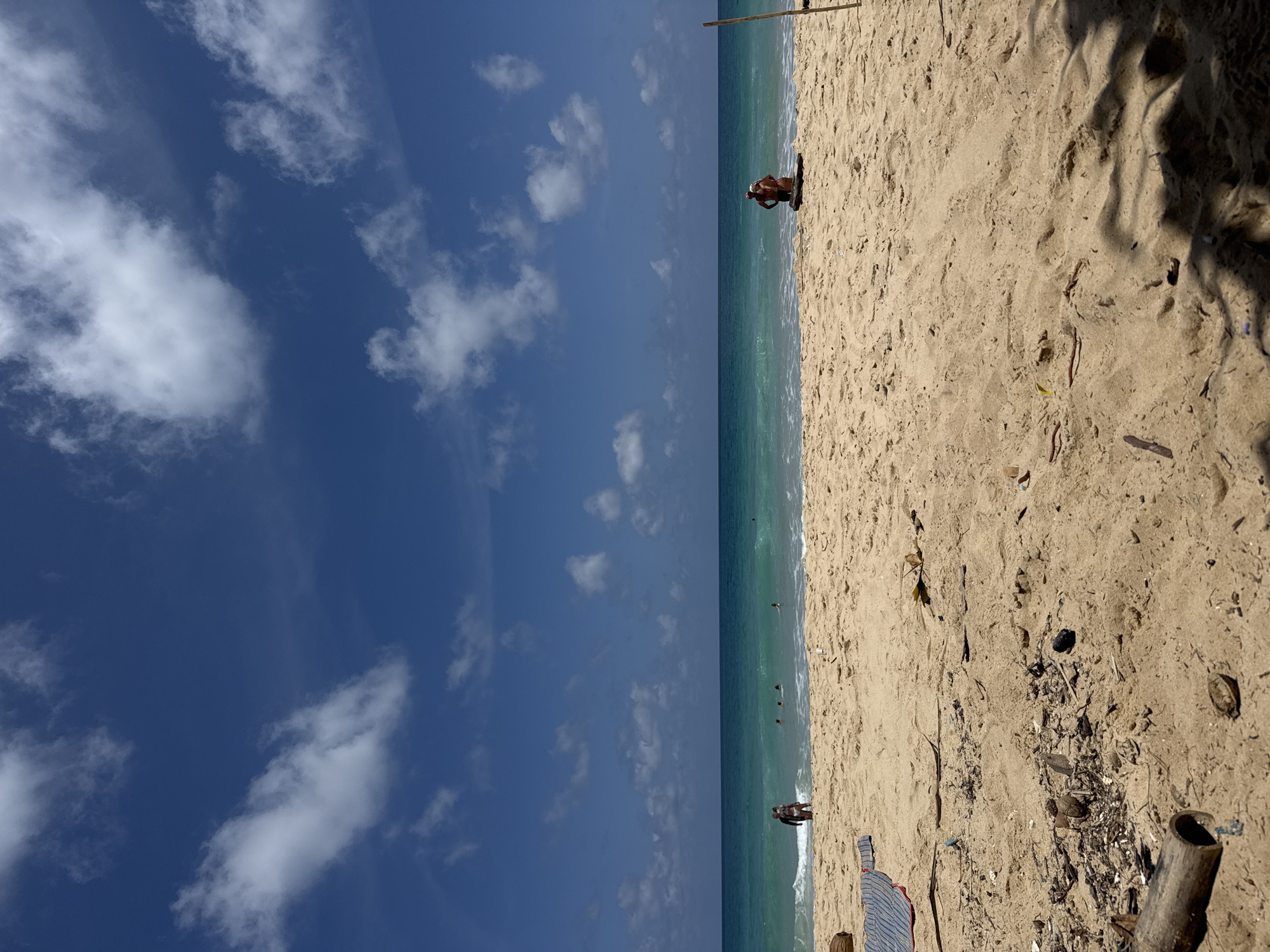 A wide-angle, low-angle, eye-level shot captures a sunlit beach scene in Choeng Thale, Thailand, under a bright blue sky dotted with fluffy white clouds. The foreground is dominated by light brown sand, strewn with scattered debris including small pieces of wood, dried leaves, and some plastic fragments. A striped towel lies on the sand to the left, and a segment of bamboo is visible in the lower left corner.
In the mid-ground, the turquoise ocean meets the sandy shore with gentle waves breaking. Several people are visible near the water's edge. On the left, two individuals stand close together, appearing to be a couple. Further out in the water, a few more people are swimming or wading. On the right side of the mid-ground, a couple is positioned near the water's edge, with one person appearing to embrace the other. They are standing near some dark driftwood. A thin wooden pole stands vertically on the far right, marking the edge of the beach. The overall atmosphere is relaxed and tropical.
