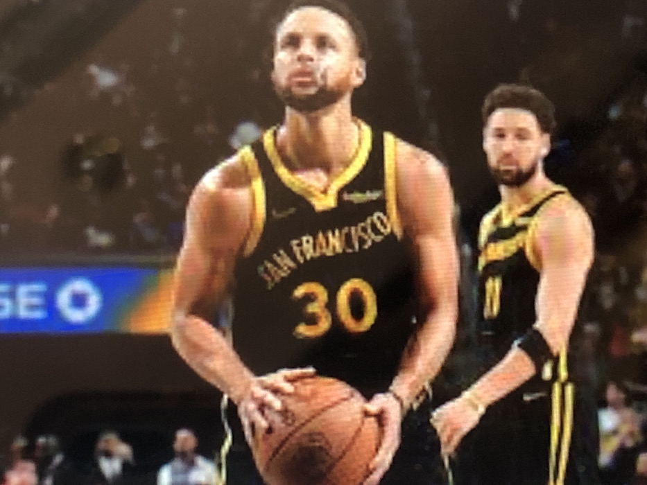 A rotated vertical image depicts two male basketball players in a professional indoor arena, situated in Kafr Sousa Municipality, Syria. In the foreground, a player wearing a black and gold sleeveless jersey with "SAN FRANCISCO" text and the number "30" holds a brown basketball with his left hand. He has short hair and a light beard. Behind him, slightly to the left, another player in a similar black and gold jersey is visible, also possessing short hair and a light beard. A black forearm band is visible on his left arm. Both individuals are looking generally to their right. The background is dark and blurred, showing minimal detail except for a vertical blue and orange structure with white lettering on the far left.