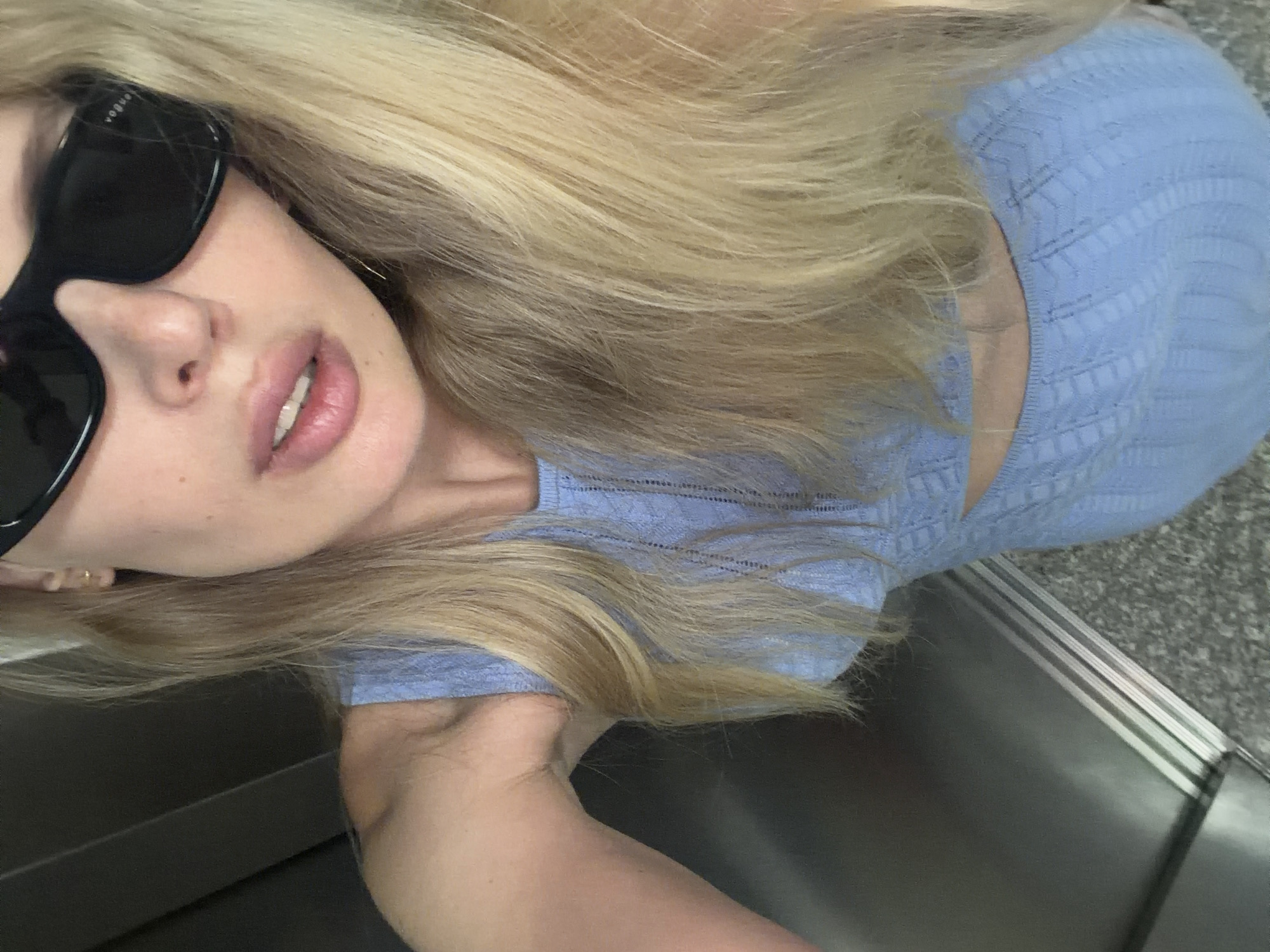 A blonde female individual is captured in a downward-angled selfie within an elevator in the city of Budva, Montenegro. Her long, straight blonde hair cascades over her shoulders. She wears large, dark, rectangular sunglasses with the brand "Vogue" visible on the right temple. A small gold-colored stud earring is visible in her right ear. Her lips are full and slightly parted, revealing a glimpse of her upper teeth. She is dressed in a light blue, form-fitting, sleeveless outfit, possibly a dress or two-piece, featuring a delicate openwork knit pattern and a cut-out section around the midriff. The setting appears to be an elevator interior, characterized by metallic, brushed silver walls and a speckled grey and white tiled or stone floor. Vertical metallic door tracks are visible where the wall meets the floor.