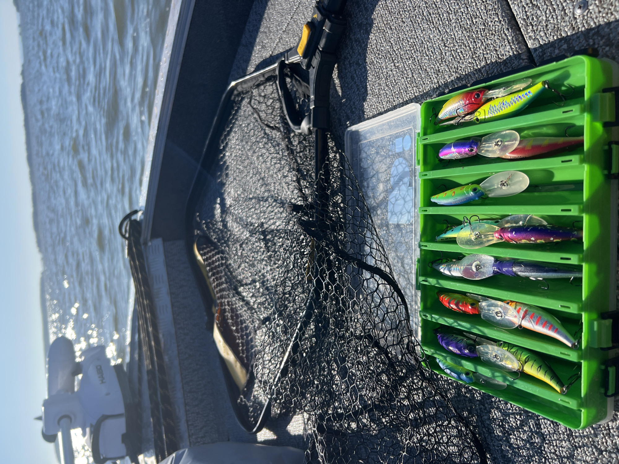 A fishing boat is on the water under a bright sky. In the foreground, a green tackle box holds various colorful fishing lures. Behind the tackle box is a black fishing net. Further back, a white electric motor with the brand "HAIBO" is visible on the boat. The water is choppy, and the sun reflects off its surface. The scene is set on a body of water, likely a lake or river, with a distant shoreline visible.