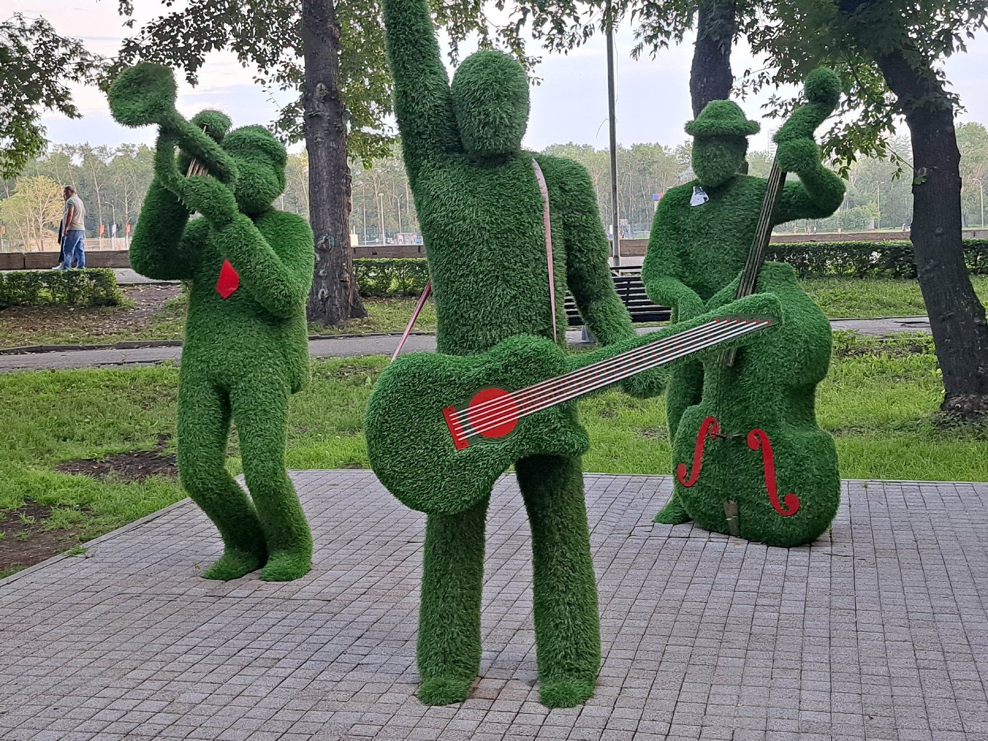 This is a full, eye-level shot of three topiary sculptures of musicians in a park, likely in Irkutsk, Russia, based on the available metadata. The figures are made of green, artificial grass. The sculpture on the left appears to be playing a trumpet. The central sculpture is larger and is playing a guitar and has its right arm raised. The sculpture on the right is playing a double bass and is wearing a hat. The figures are on a paved walkway made of light gray square tiles. Behind the sculptures, there is grass and trees. In the background, there are more trees and a few people walking. The sky is overcast.