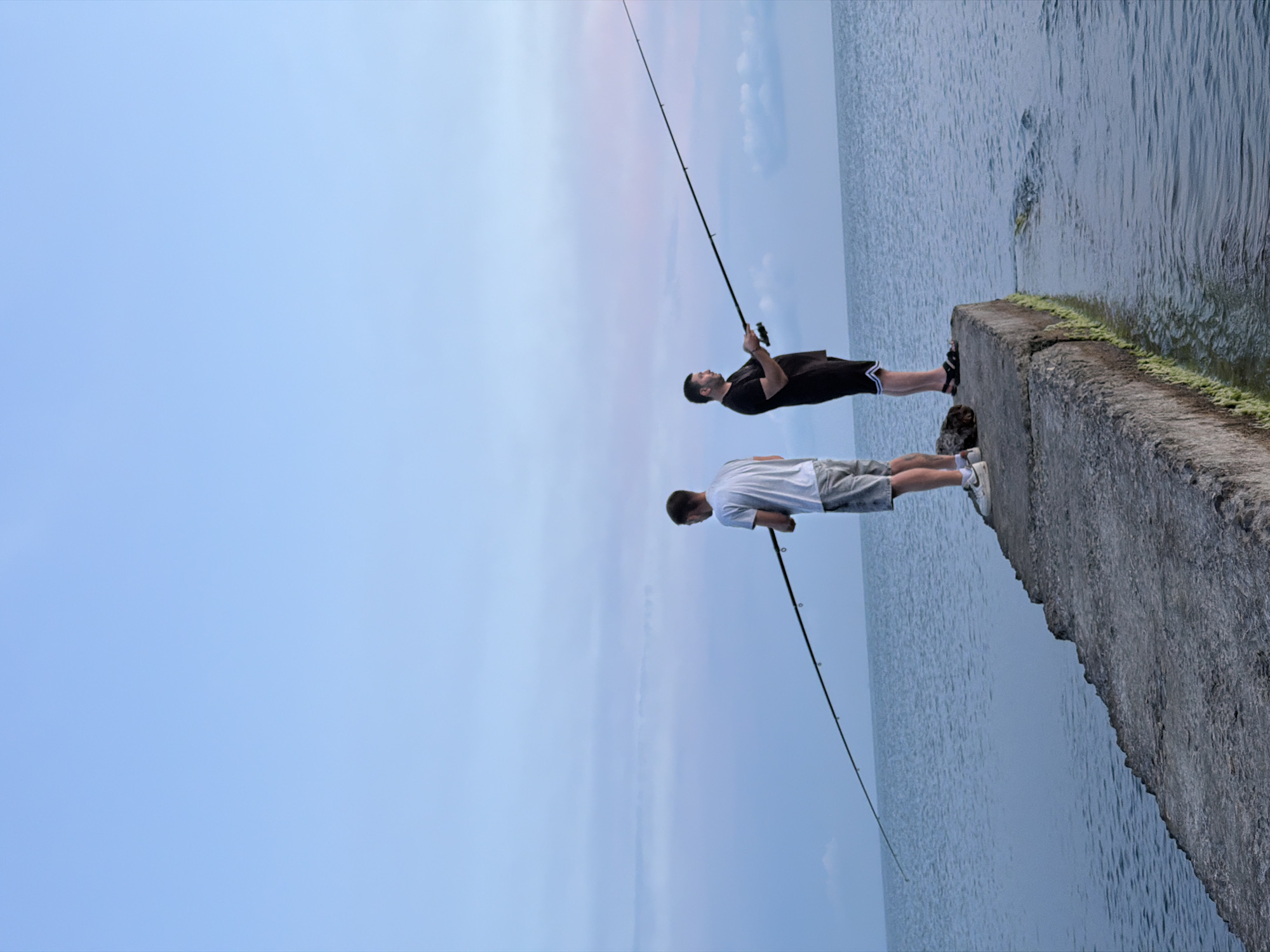 Two men are fishing on a concrete pier at Dederkoy, Russia. The man on the left is wearing a white t-shirt and shorts, and the man on the right is wearing a black t-shirt and black shorts. Both men are holding fishing rods. The ocean is calm, and the sky is a pale blue with some clouds. The pier is weathered and has some green algae growing on its sides. The image captures a serene moment of recreational activity by the sea.