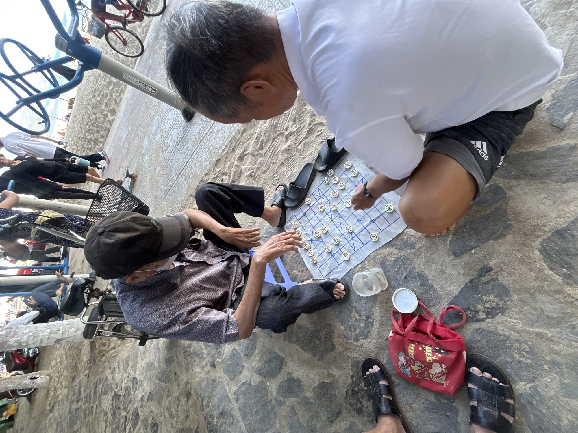 Two older men are playing a game of Chinese chess on a makeshift board on the stone ground in the city of Bac Nha Trang Ward, Vietnam. One man is kneeling, wearing a white shirt and gray shorts with an Adidas logo. The other man is sitting on a small blue stool, wearing a cap, face mask, and patterned shirt. They are surrounded by sandals and a red bag with illustrations. In the background, there are exercise machines, bicycles, and other people.