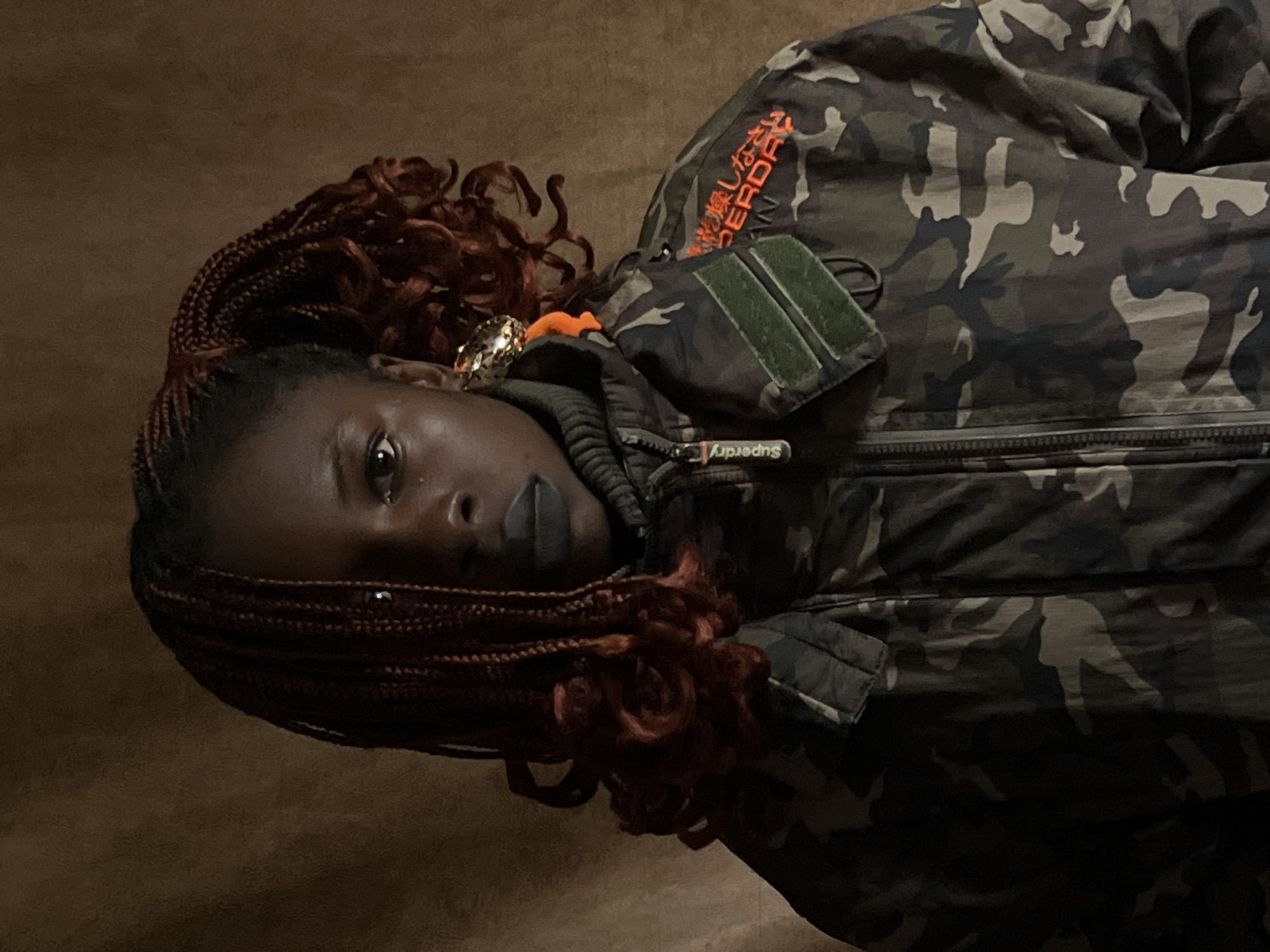 A portrait of a young Black woman with dark skin and braided hair, wearing a camouflage jacket over a dark turtleneck. She has dark lipstick and a large, ornate earring in her right ear. The background is a textured, brown fabric. The image is cropped to focus on the woman's upper body and face. The lighting is dim and casts shadows, creating a moody atmosphere. The woman's gaze is directed towards the camera. There is a patch on the jacket with orange text that appears to be in Japanese. The setting does not provide any specific indicators of Jos, Nigeria.