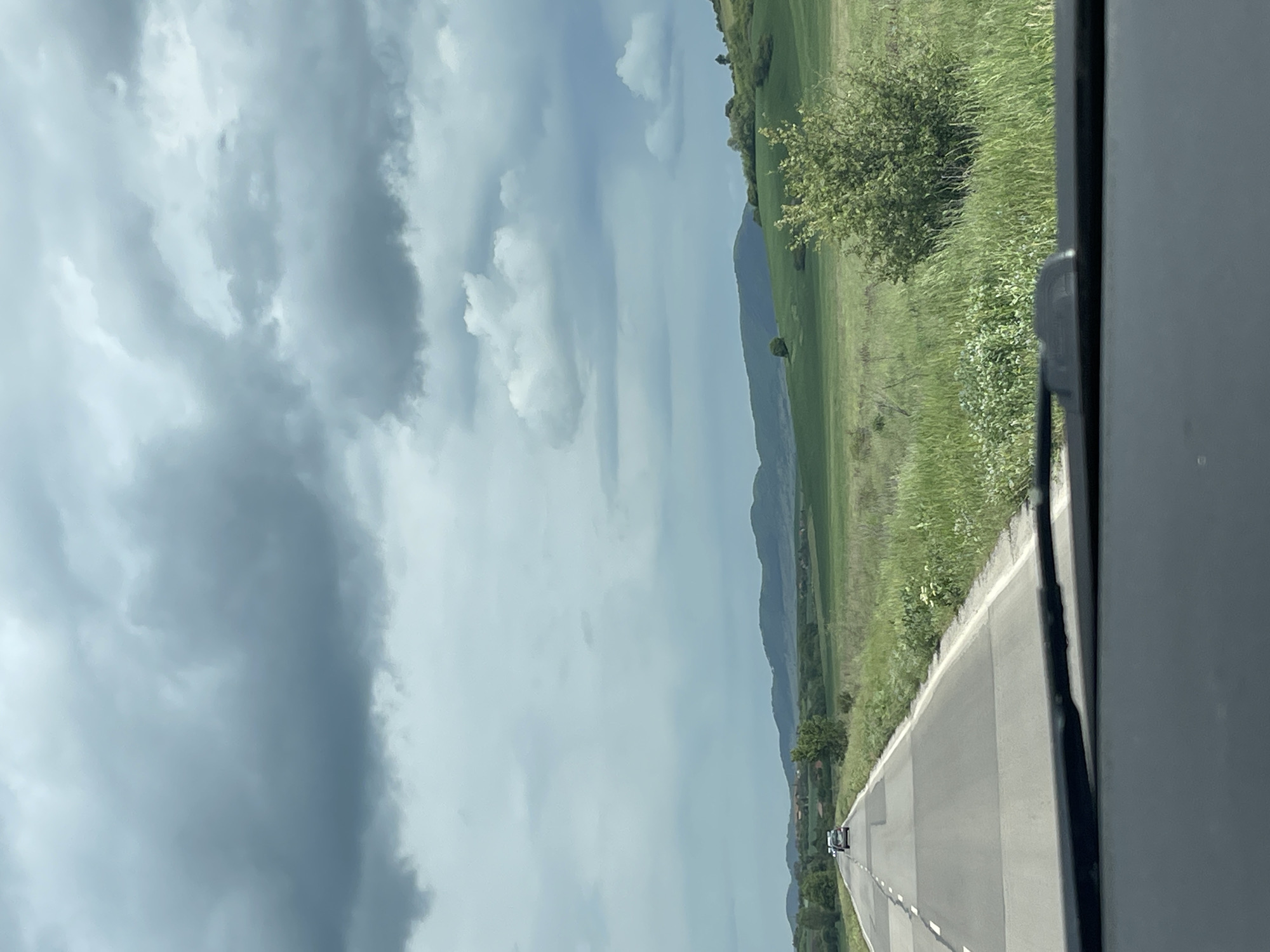A horizontal landscape photograph taken from inside a vehicle shows a road receding into the distance in Batanovtsi, Bulgaria. The sky, occupying the upper portion of the frame, is filled with gray and white clouds. To the right of the road is a field of green grass and scattered trees that extends towards a distant mountain range. The asphalt road has white lane markings and is bordered by a concrete curb. A car can be seen on the road in the distance. The edge of the vehicle's interior is visible on the right side of the frame.