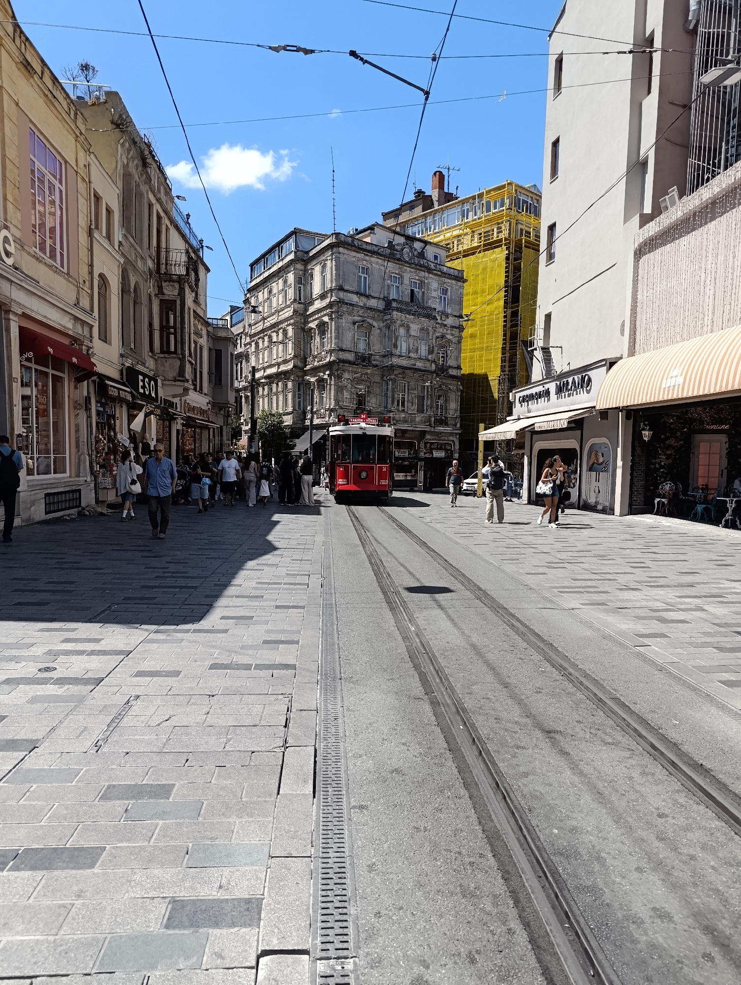 In Beyoğlu, Turkey, a bright, sunny day is captured on a street with tram tracks running down the center. The street is paved with grey rectangular stones, and on either side, buildings of various architectural styles line the thoroughfare. On the left, a yellow and beige building with ornate windows and balconies is visible, with awnings extending from some of the shops. Further down, a vintage red tram car with the number 31 on its front is stopped on the tracks, its doors open.
On the right side of the street, a lighter colored building with a modern facade stands tall, featuring large windows and a striped awning. A few people are walking on the sidewalk, some browsing shop windows, while others are in transit. The sky is a clear blue with a few wisps of white clouds. The scene depicts a bustling urban environment with a blend of old and new architecture, typical of a vibrant city street.