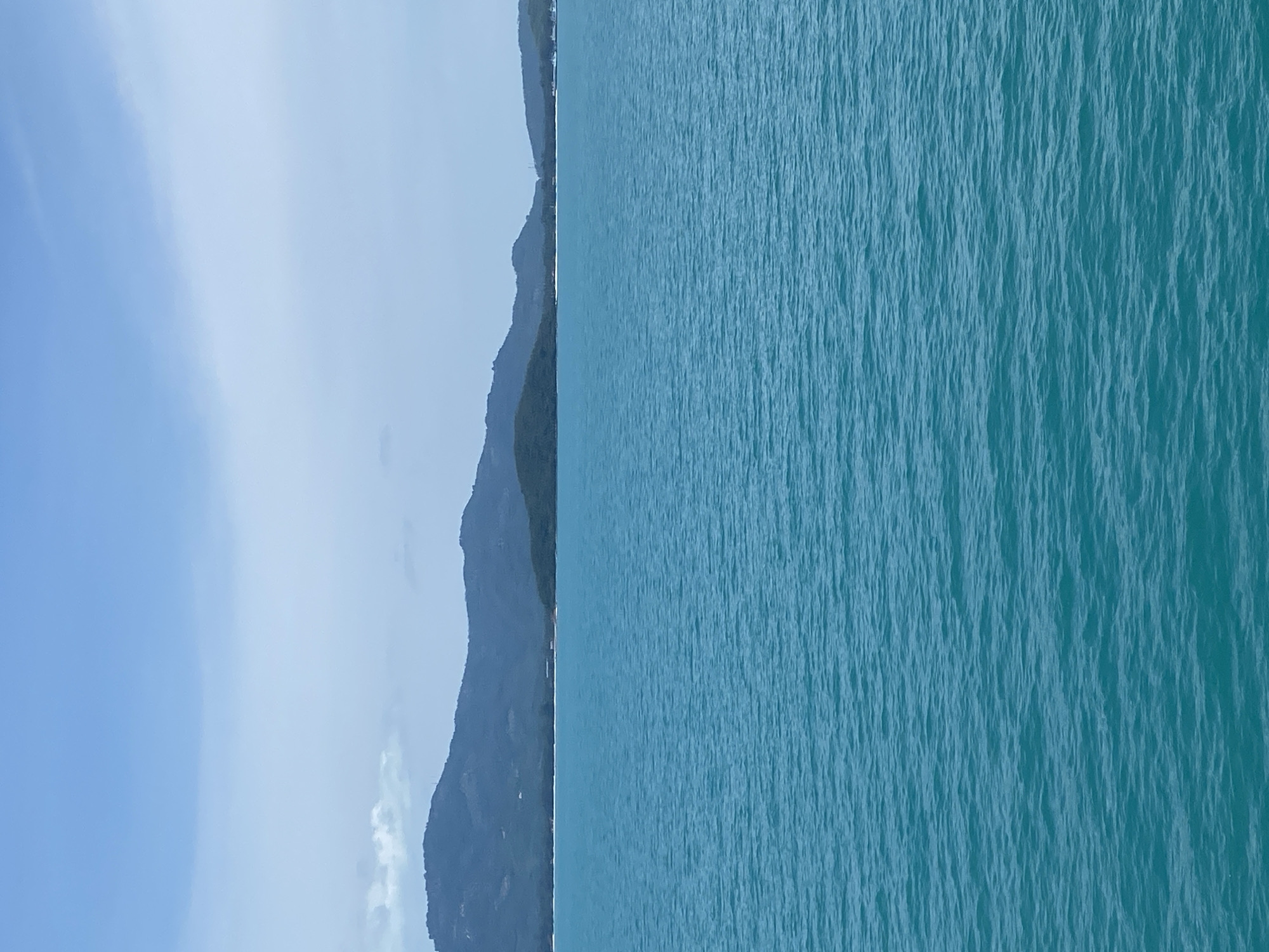 A wide, vertical shot depicts a serene turquoise ocean under a pale blue, slightly overcast sky. In the distance, a chain of lush green, undulating hills forms a coastline, their slopes dotted with dark vegetation. A thin, pale sand beach is visible along the shoreline, separating the water from the hills. The ocean surface is textured with gentle ripples and waves, creating a dynamic yet calming visual. The overall scene evokes a sense of tranquility and natural beauty, likely a coastal landscape in Thailand, given the prominent turquoise waters and tropical-looking hills.