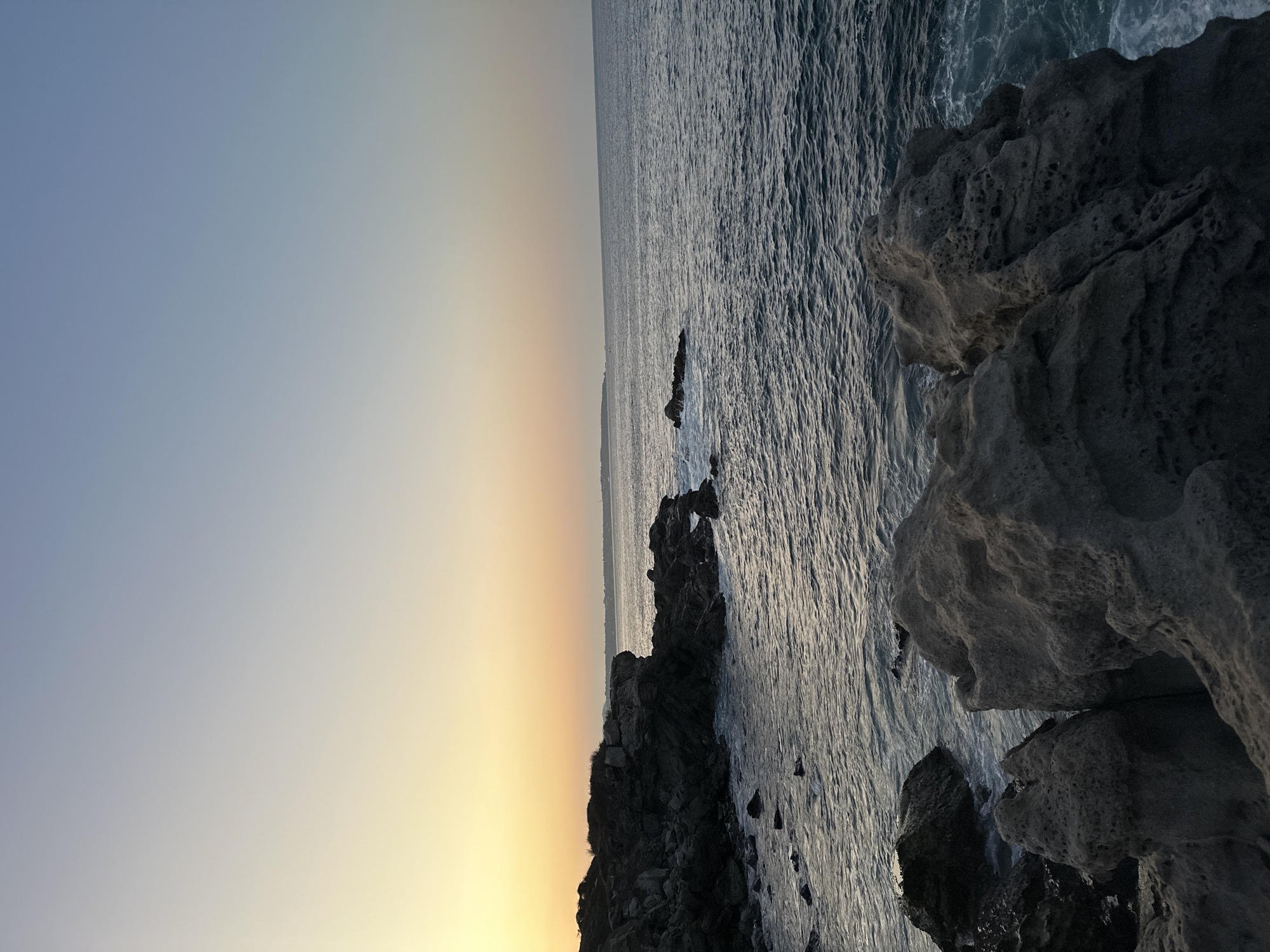 The image shows a rocky coastline meeting the ocean at sunset. The sky fades from light blue to a golden yellow near the horizon. The ocean is dark and textured with small waves. Jagged, dark-colored rocks line the right side of the image. The location is likely near Puerto Escondido, Mexico.