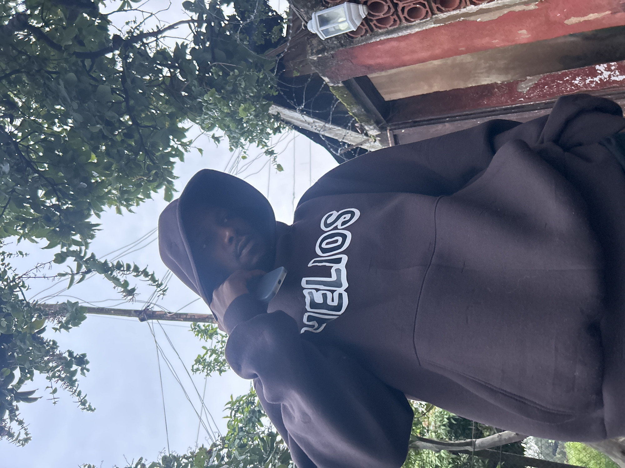 A low-angle, medium shot captures a person wearing a dark brown hooded sweatshirt with the word "VELIOS" emblazoned in white on the chest. The person is holding a smartphone to their ear with their right hand, partially obscuring their face. They are standing outdoors, with lush green trees and branches overhead. In the background, a weathered, reddish-brown wall with intricate circular patterns and barbed wire can be seen, suggesting a potentially industrial or older urban setting. A white outdoor lamp is mounted on the wall. The overall environment appears to be an outdoor space within a city, possibly residential or commercial, with elements of both nature and urban infrastructure. The sky is overcast and light-colored. This scene could plausibly be located in Jos, Nigeria, given the visual context of vegetation, building style, and typical urban elements.