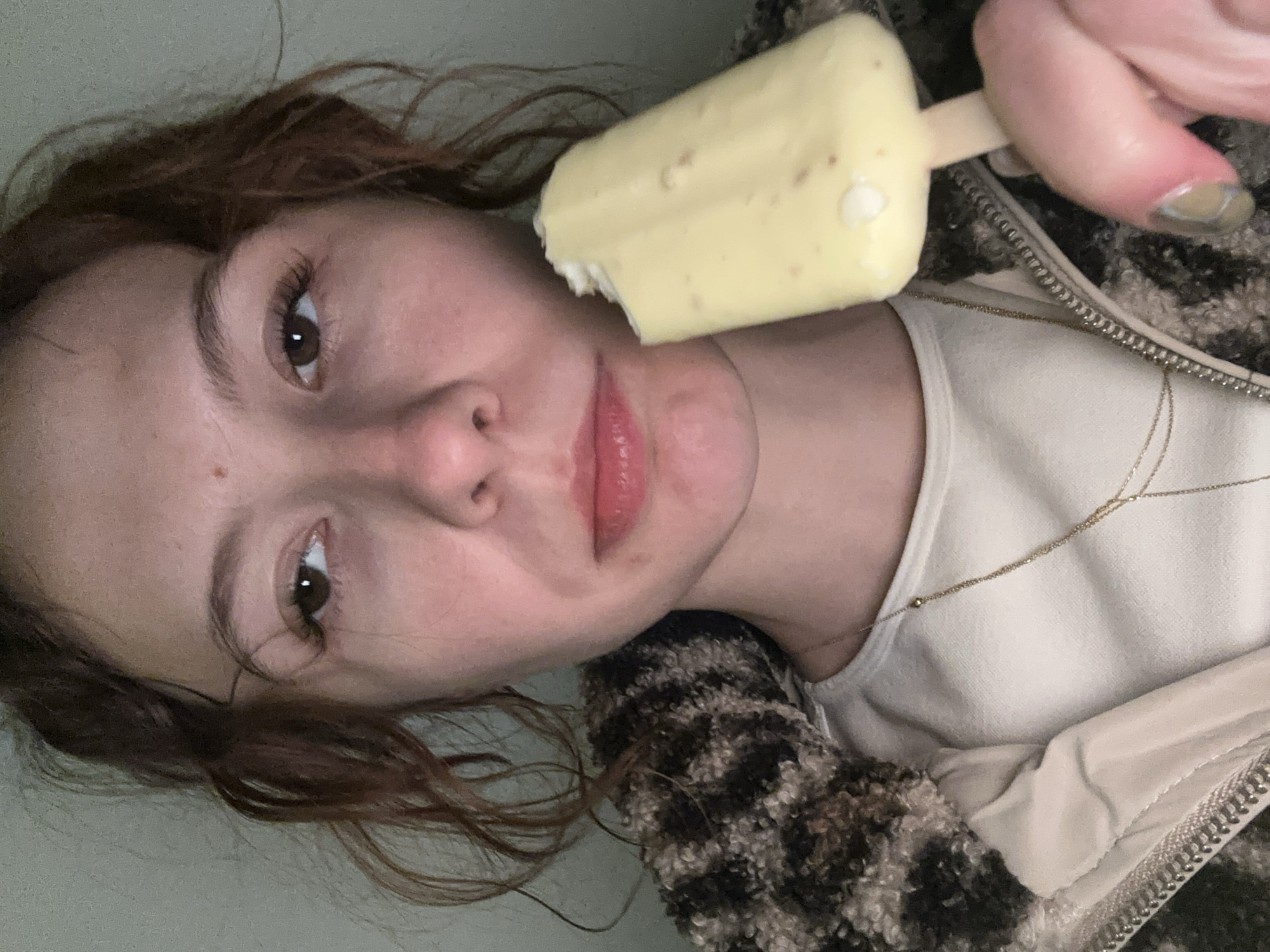 A close-up, rotated portrait shows a young person in Novi Sad, Serbia, holding a light yellow popsicle or ice cream bar with a bite taken out of it. The subject's face is prominent, featuring dark brown eyes that look directly at the viewer, framed by long eyelashes. Their lips are a soft pink, slightly parted, and their expression is largely neutral, possibly pensive.
The person has brown, wavy hair and is wearing a light cream or off-white top, over which a textured jacket with a dark, mottled, possibly animal print pattern is visible. A thin, gold-colored chain necklace with small accents rests on their chest. The hand holding the popsicle shows nails with traces of polish. The background is a plain, light-colored surface, indicating an indoor setting. There is no visible text in the image. The lighting is soft and ambient.