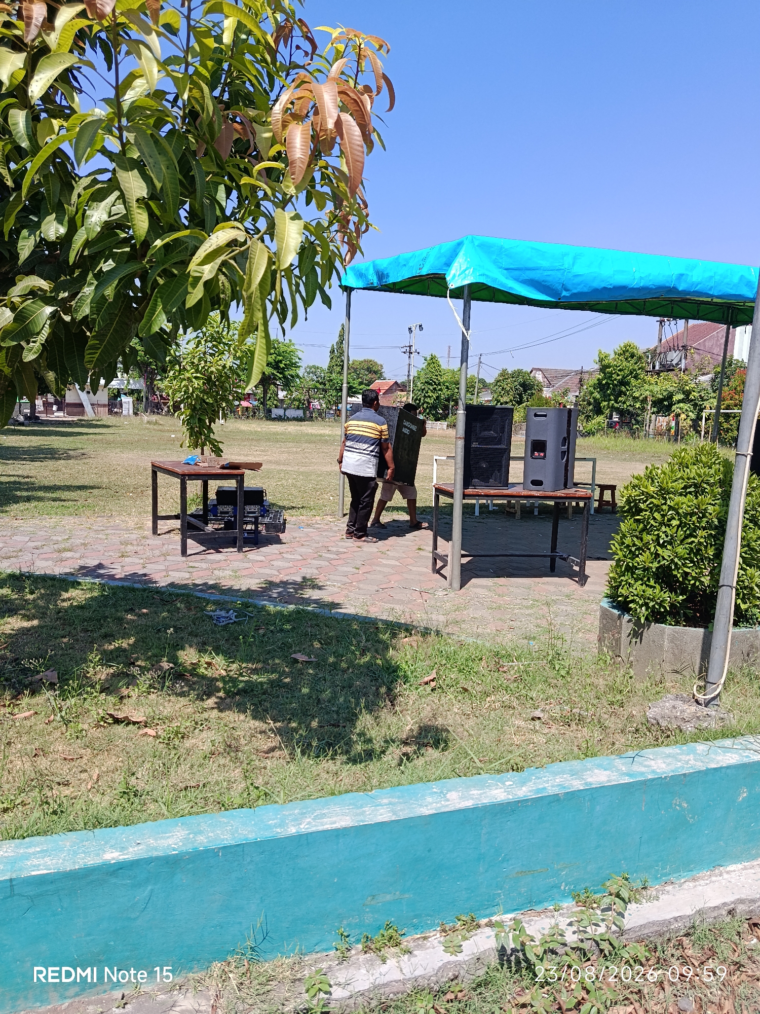 The image depicts an outdoor scene in Keboansikep, Indonesia, on August 23, 2020, at 09:59. Two men are present near a portable blue canopy set up on a grassy area with a paved walkway. One man, wearing a striped shirt and dark pants, stands by a table that holds two large black speakers and other audio equipment. The second man is partially visible, carrying what appears to be a speaker or a piece of audio equipment. In the foreground, a low, turquoise-painted wall runs across the bottom of the frame, with grass and some dry leaves on the ground. To the left, a large tree with green and reddish leaves casts a shadow. In the background, there are more trees, houses, and utility poles, suggesting a suburban or semi-rural environment. The sky is clear and blue.