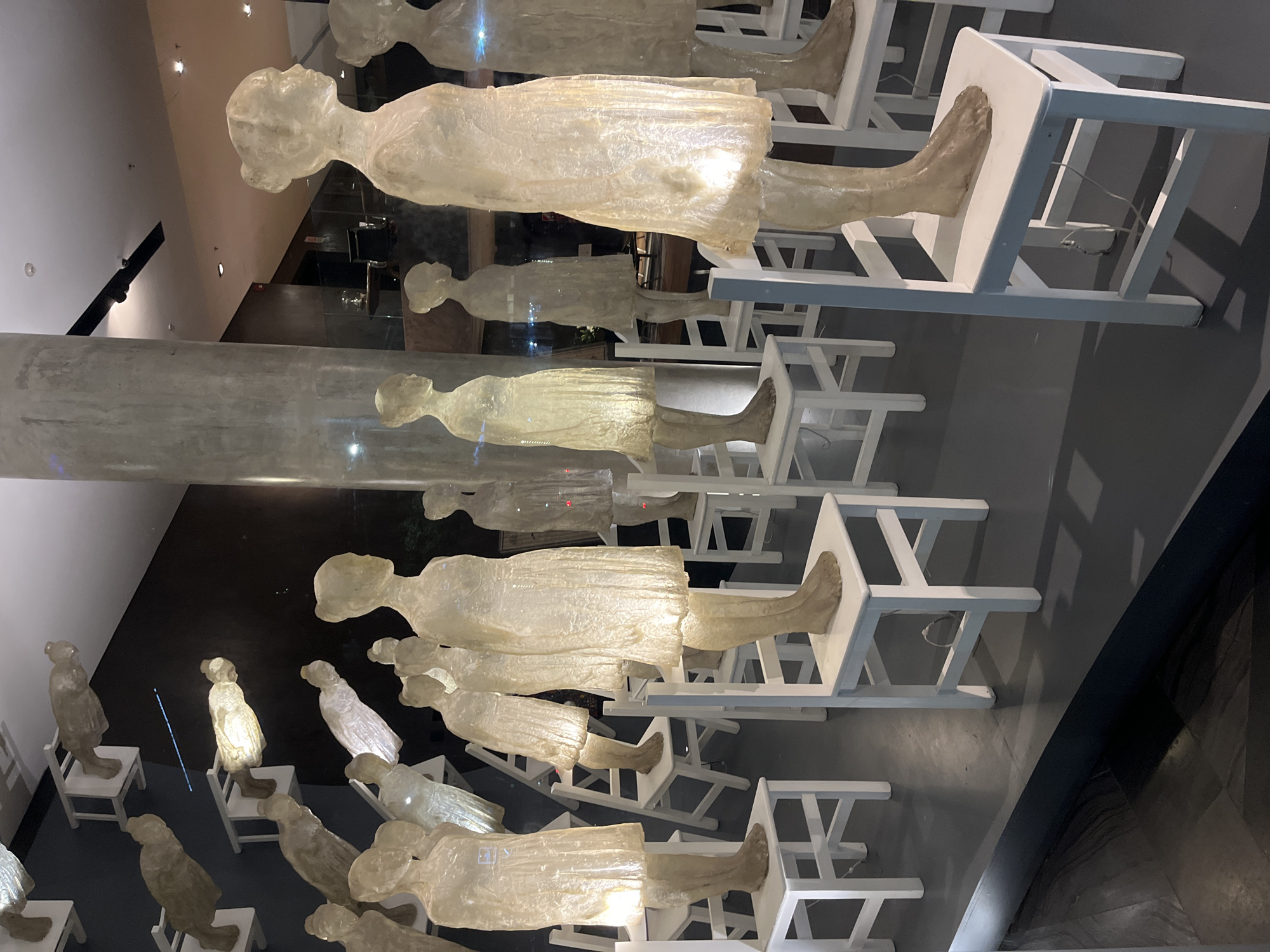 The image shows an art installation in what appears to be a gallery or museum in Buenos Aires, Argentina. The installation consists of numerous translucent, possibly resin or glass, human figures positioned sitting on white chairs. The figures are arranged in a tiered or cascading formation. The figures are vaguely humanoid in shape, with simple outlines of heads, bodies, and limbs. The figures are subtly illuminated from within, some more brightly than others. The chairs are simple white wooden frames with flat seats. The floor is tiled with dark tiles. In the background, some architectural features like walls, ceilings and a support are visible. There are other unidentifiable objects or reflections visible on the left.
