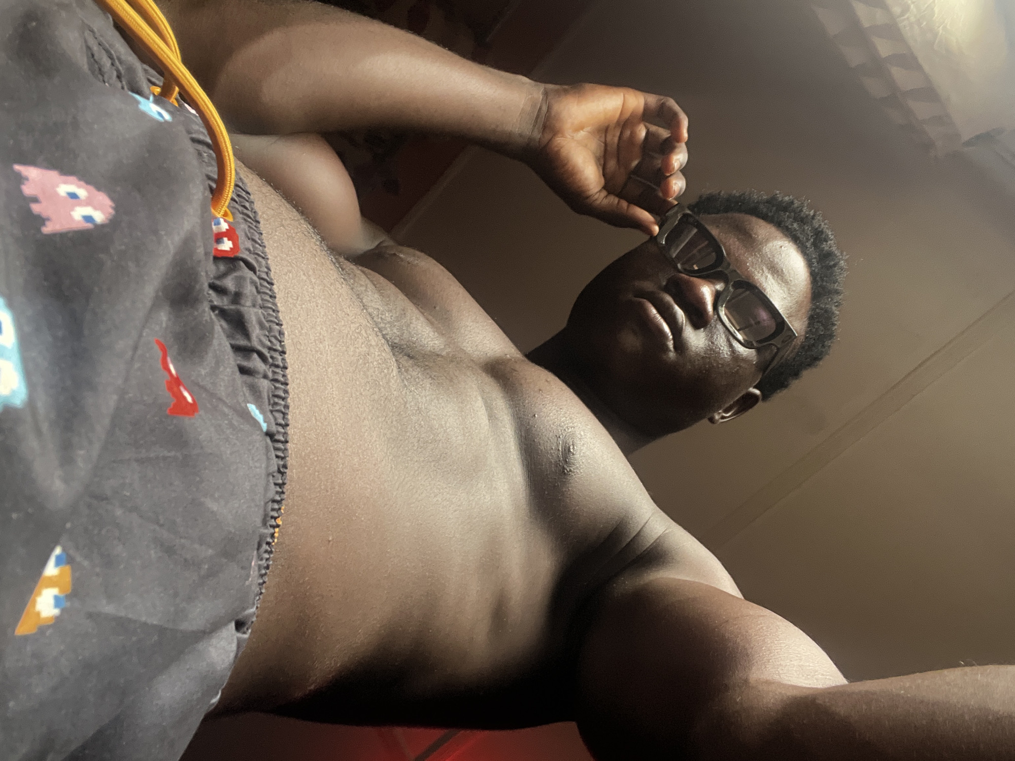 A shirtless, dark-skinned man is depicted from a low angle. He is wearing black glasses and dark shorts with a Pac-Man pattern. His right hand is lifting the glasses slightly off his face. The background is out of focus, showing indistinct walls and ceiling. There is no discernible evidence to place the scene in Mosafejo, Nigeria.