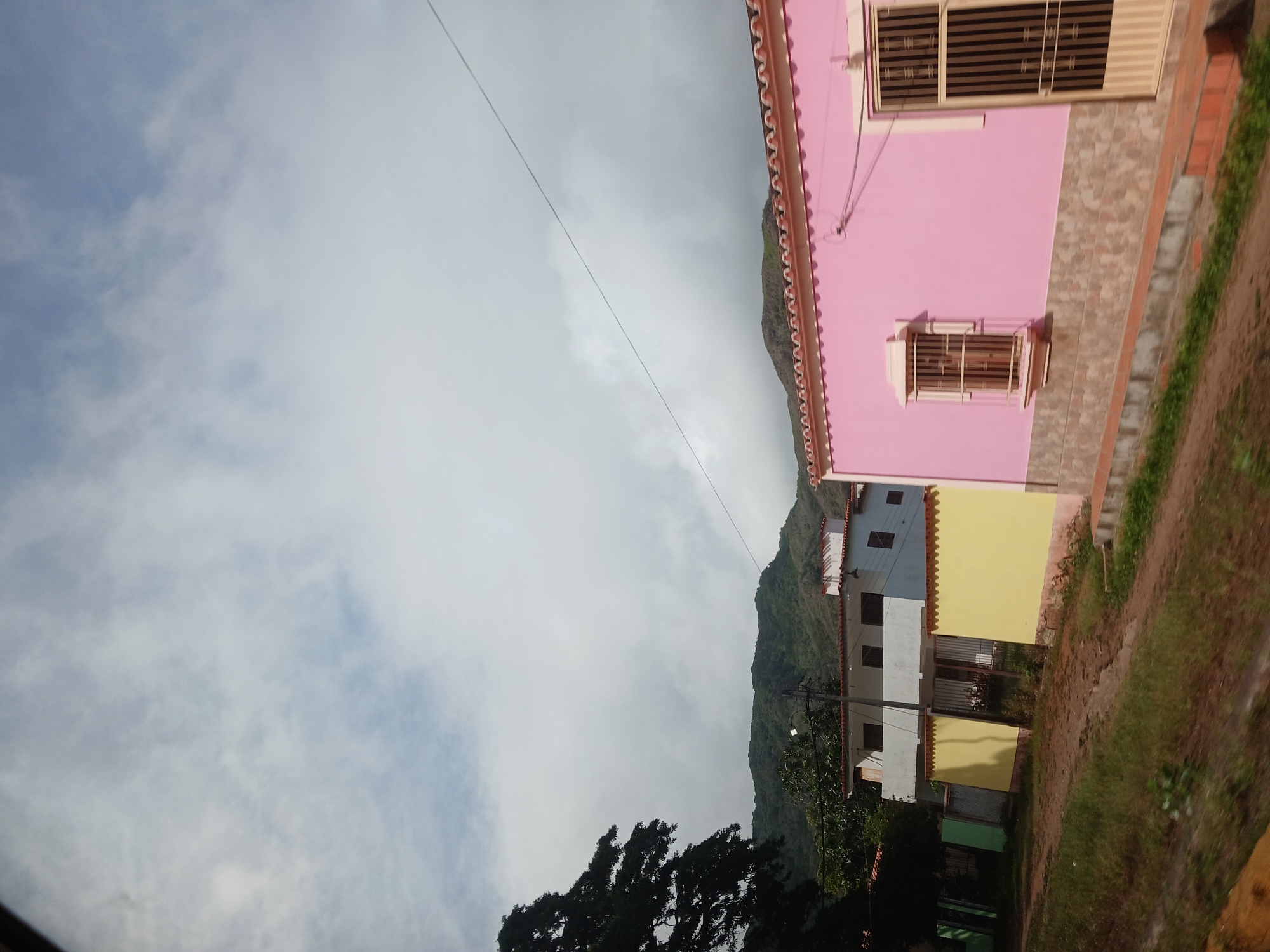 This is a medium-shot, outdoor, daytime photograph of a residential street in Tacarigua, Venezuela. The foreground shows a dirt road with dry grass and some green vegetation. To the right, a pink-colored house with a tiled roof is visible. This house features a window with decorative iron bars and a beige door with vertical bars. The lower part of the pink house is adorned with stone-like tiles. Behind the pink house, a yellow wall and a white building with a tiled roof are partially visible. This white building appears to have multiple windows. Further in the background, lush green mountains are shrouded in clouds. The sky is mostly cloudy with patches of blue. A dark-leaved tree is present on the left side of the image. A thin electrical wire stretches diagonally across the upper portion of the frame. There are no people or animals visible in the scene.