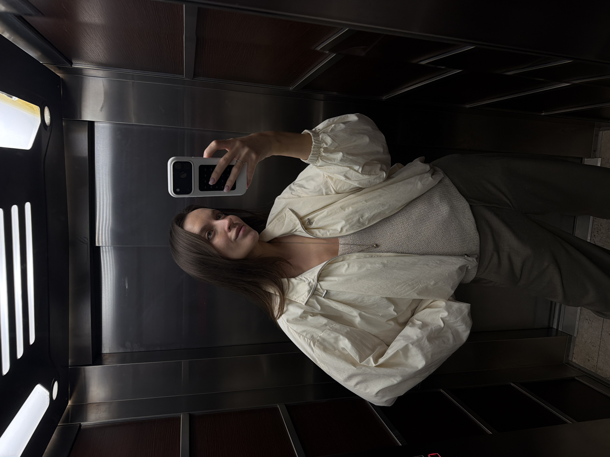 A young woman is captured taking a mirror selfie inside an elevator. She has long, straight dark brown hair and looks directly at the camera with a calm, subtle smile. She is wearing a light cream or off-white zip-up jacket with voluminous, cuffed sleeves over a fitted ribbed knit top of a similar light hue. A thin silver necklace with a small pendant is visible around her neck. In her right hand, she holds a smartphone with a white case, its screen reflecting her image, and her fingernails are painted with a light-colored polish.
The elevator interior features highly reflective metallic walls, which serve as the mirror for the selfie. These walls are bordered by darker, possibly wood-grained or textured panels at the top and bottom of the frame. On the left side of the frame, a vertical light fixture composed of several illuminated bars provides ambient lighting within the elevator. The overall lighting is somewhat dim, with the metallic surfaces reflecting the available light. The image is oriented sideways, giving the impression that the subject is lying down. No discernible text is visible on the phone or within the elevator.