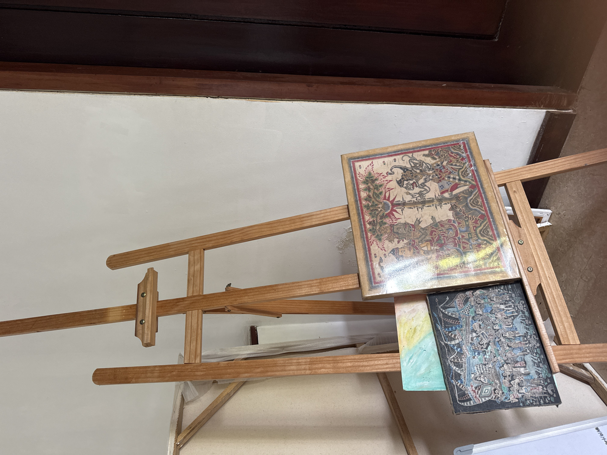 The scene captures an easel displaying two framed artworks, positioned against a white wall under a dark wooden frame or trim. The top artwork features intricate, reddish-brown detailed drawings. Underneath, the second artwork has a dark, possibly black, background with blue and gray detailed drawings. The easel itself is made of light-colored wood, suggesting a workshop or studio setting. The image was probably taken indoors in Kuta Selatan, Indonesia.